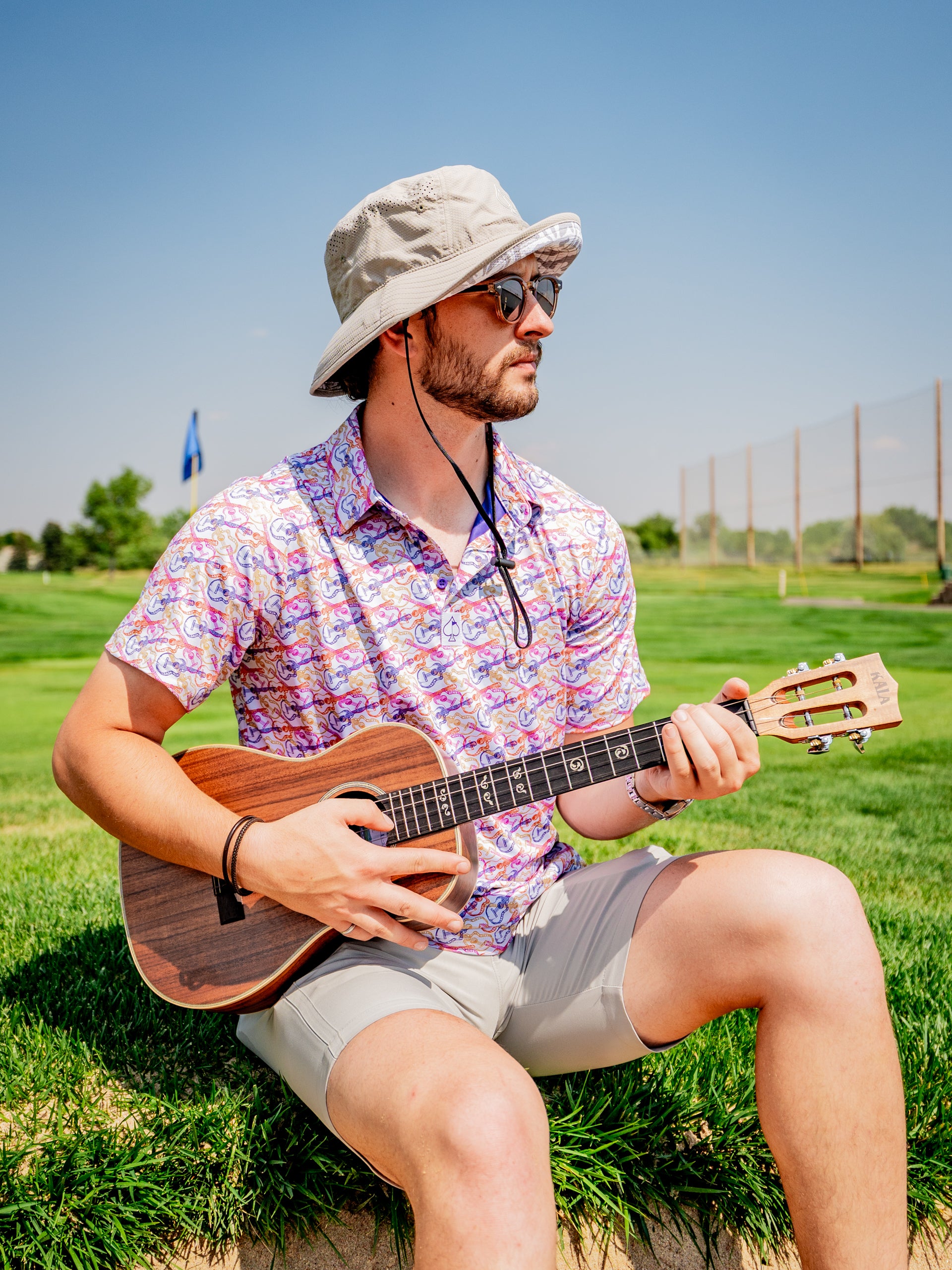 Man wearing a colorful Margaritaville shirt and bucket hat sits on grass playing a ukulele on a sunny golf course.