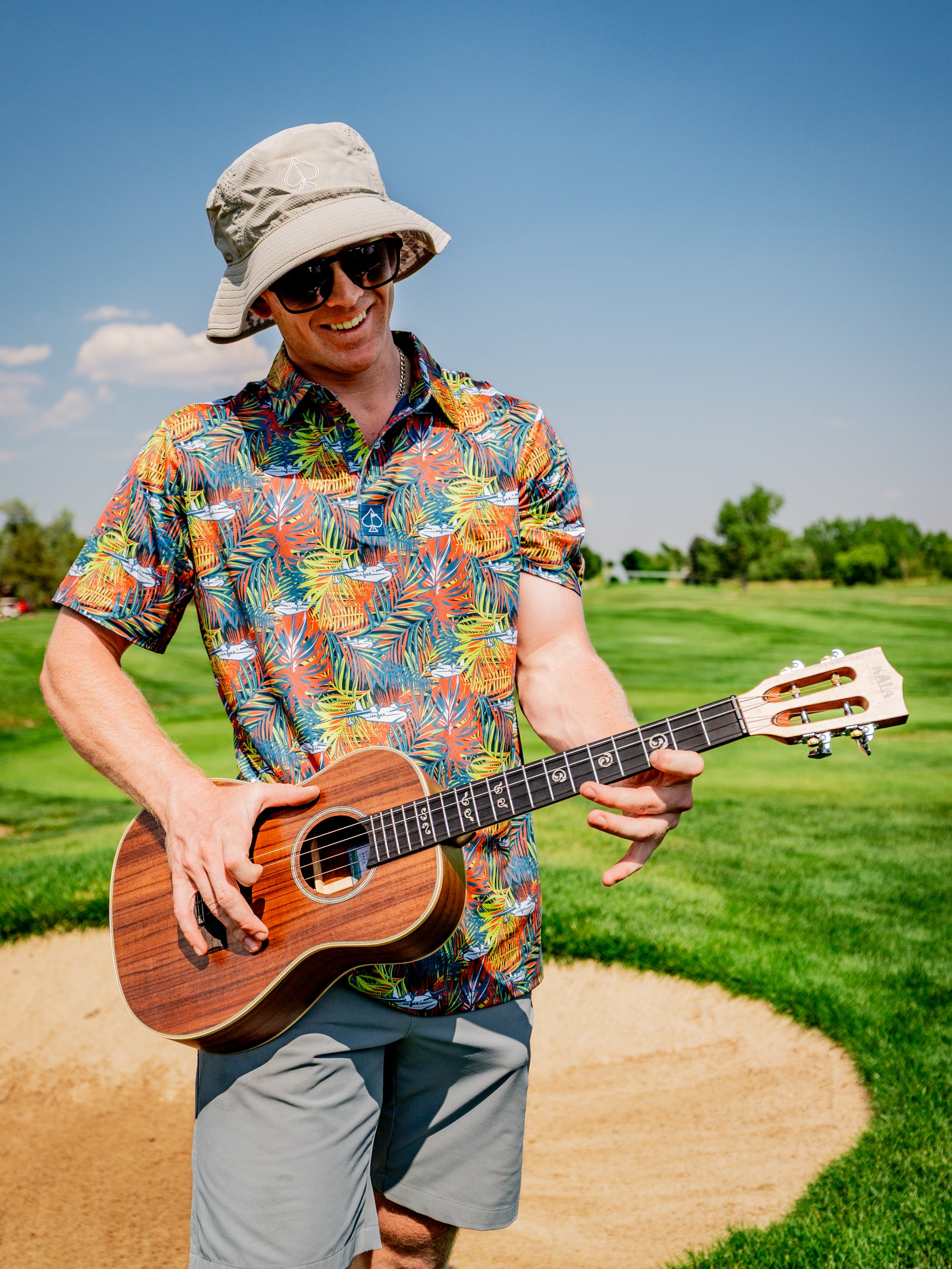 A man in a colorful tropical shirt and bucket hat plays a ukulele on a golf course, enjoying a sunny day.