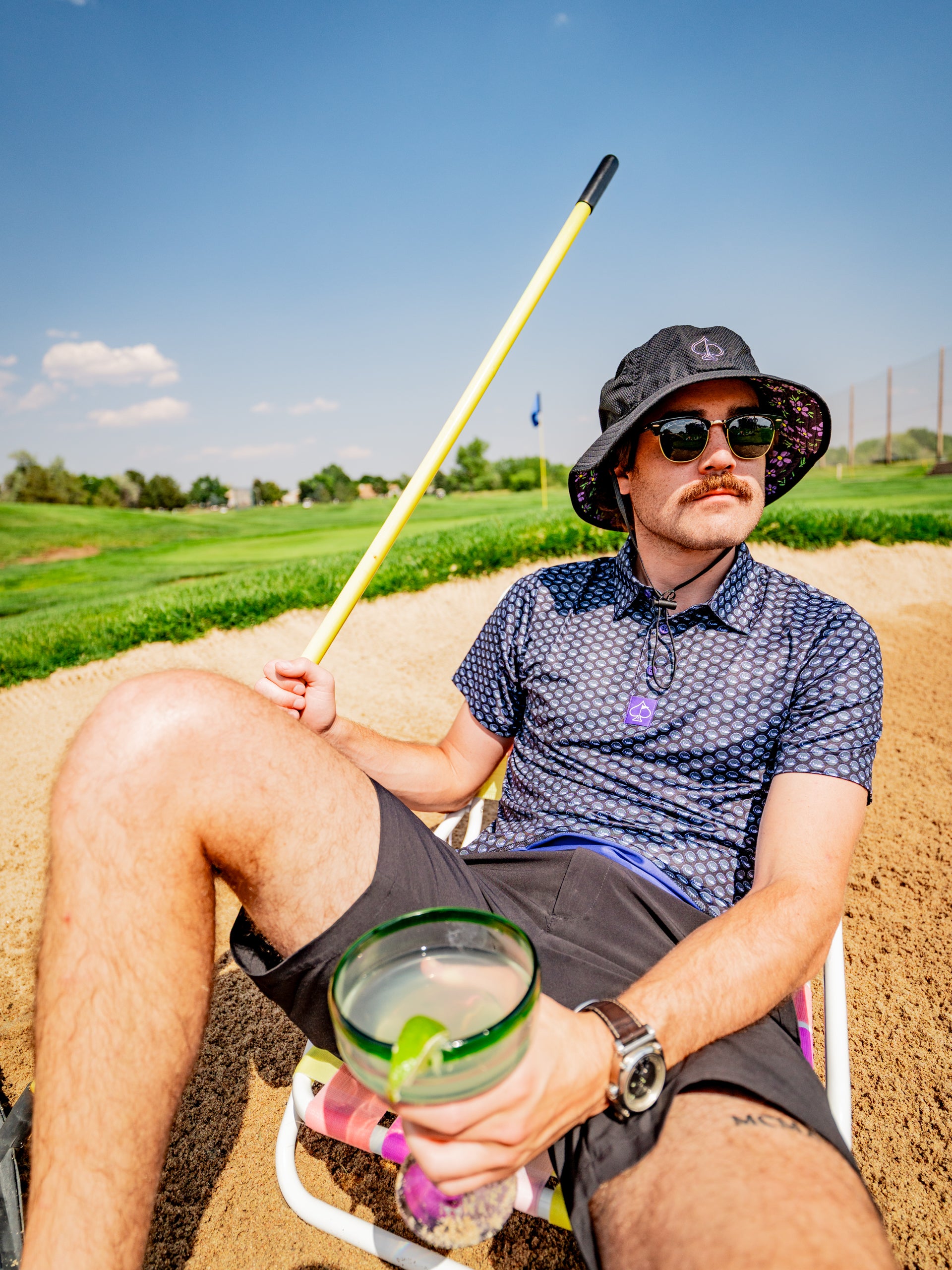 Man lounging in a golf sand trap, wearing a stylish golf shirt and bucket hat, holding a drink and a golf club.