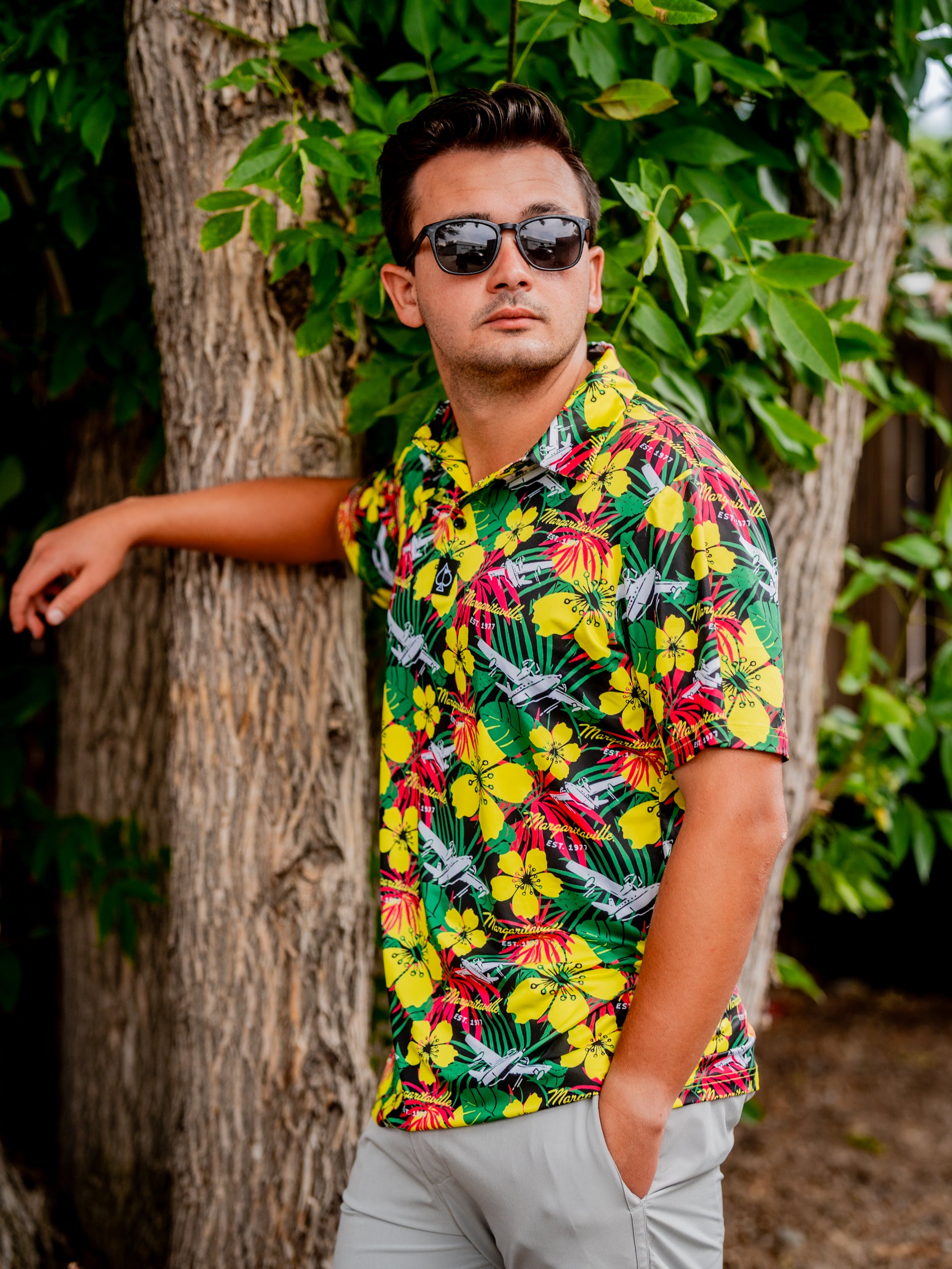 A man poses confidently in a vibrant floral polo shirt against a tree, showcasing a tropical style perfect for summer outings.