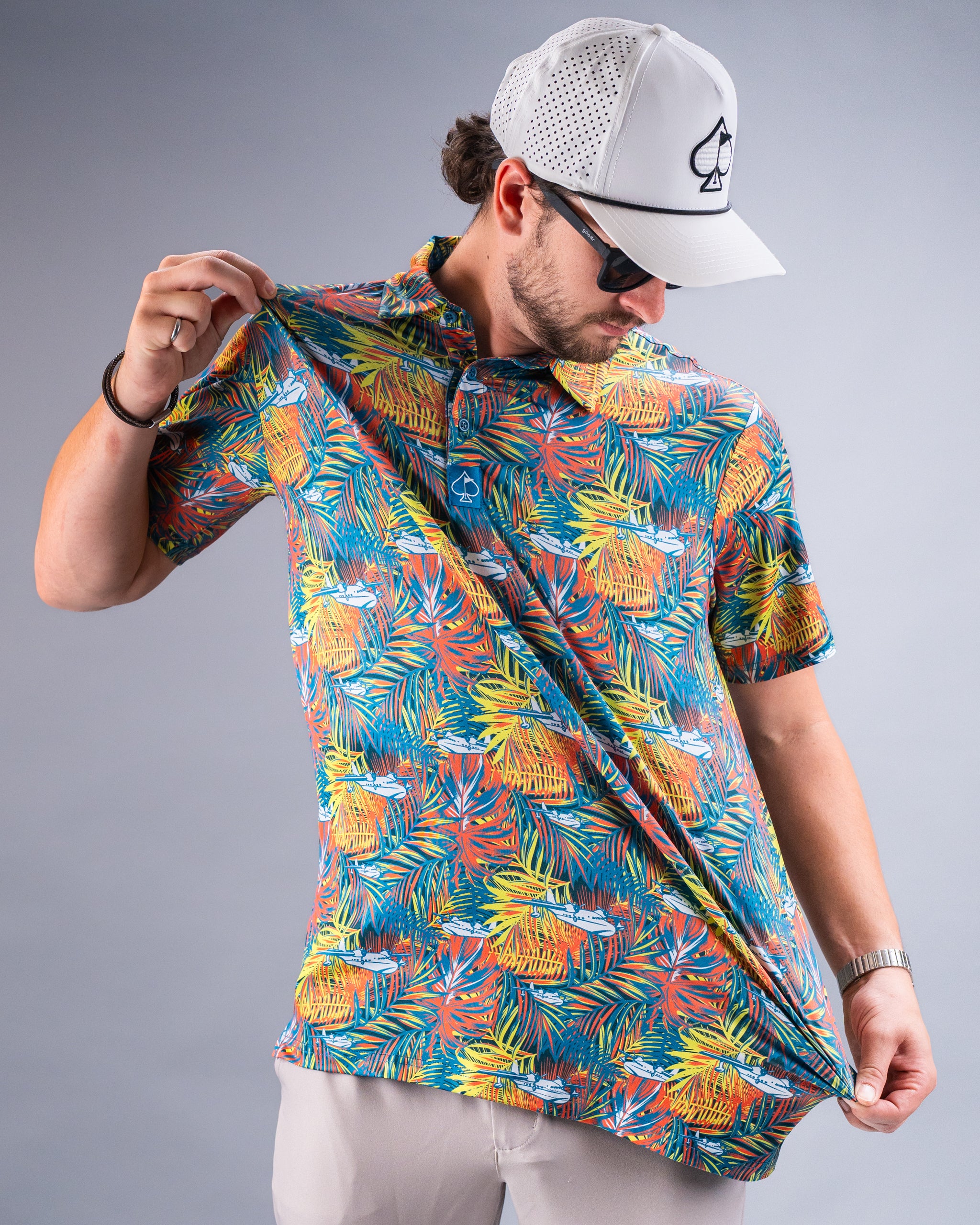 Man wearing a colorful, tropical-themed golf shirt with palm leaves and planes, paired with a white mesh cap with a spade ...