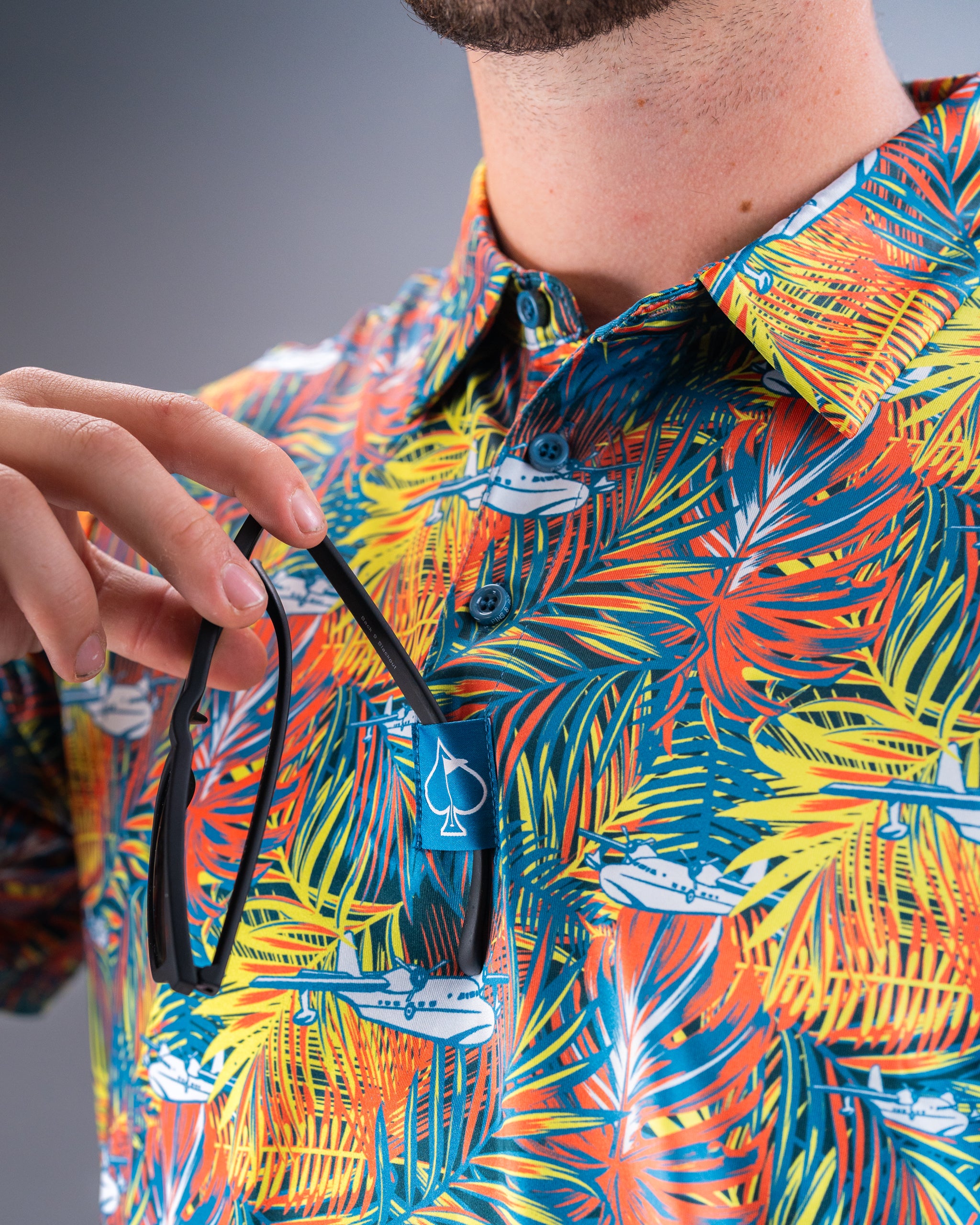 Colorful tropical print golf shirt with bold leaf and airplane patterns, featuring a blue spade logo tag. Perfect for stan...