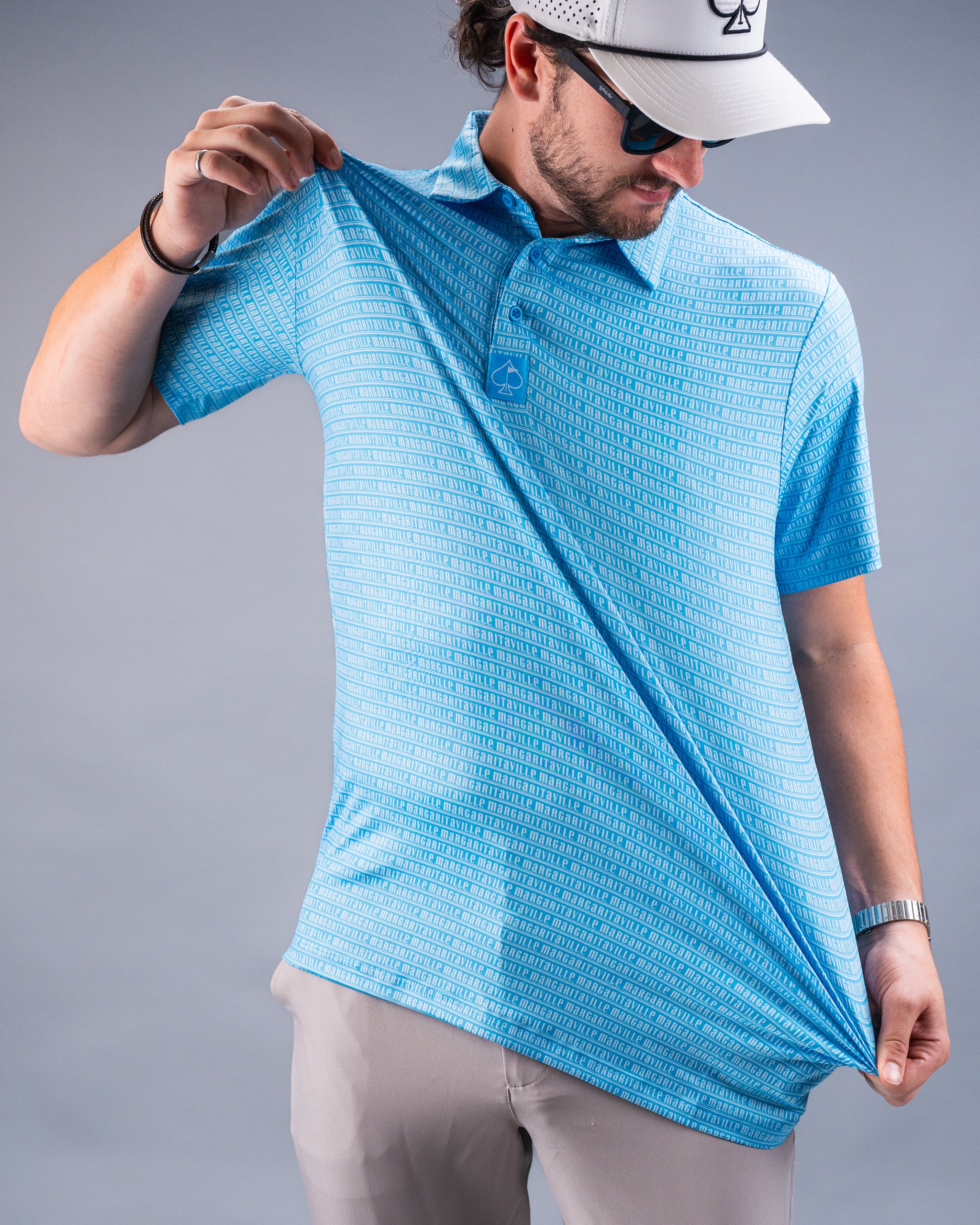 Man in blue golf polo with "margaritaville" text pattern, wearing sunglasses and a white cap, holding the shirt's hem.