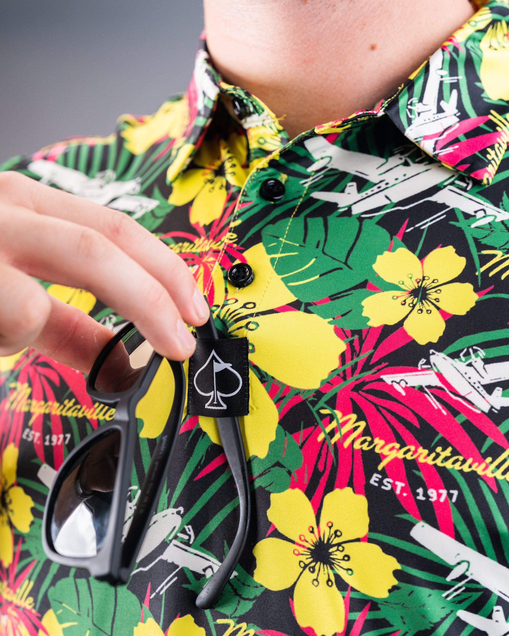 Close-up of a colorful, tropical-print golf shirt featuring yellow hibiscus flowers, green leaves, and a black tag with a ...