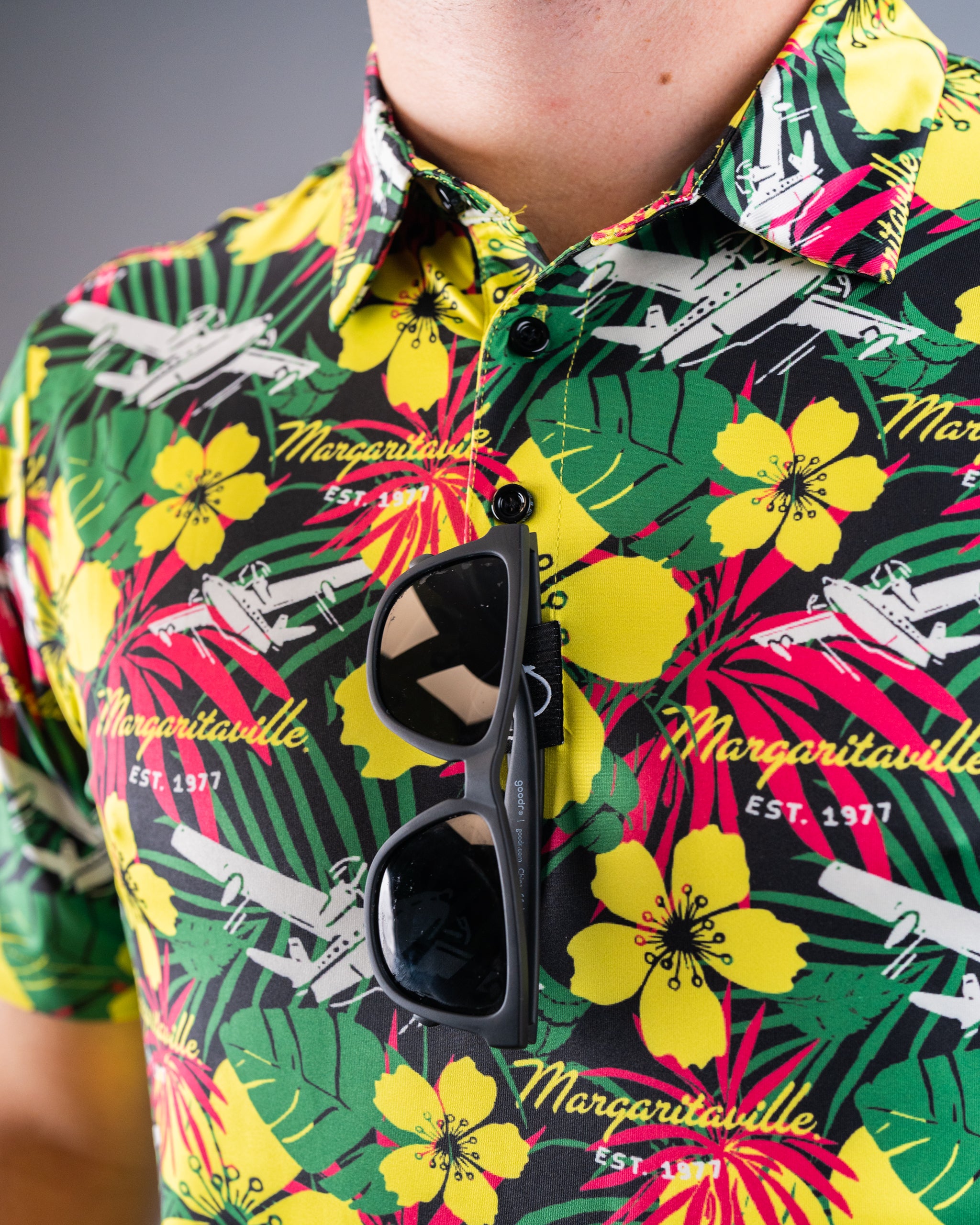 Vibrant tropical shirt with yellow hibiscus flowers, green leaves, and "Margaritaville" text; black sunglasses clipped to ...
