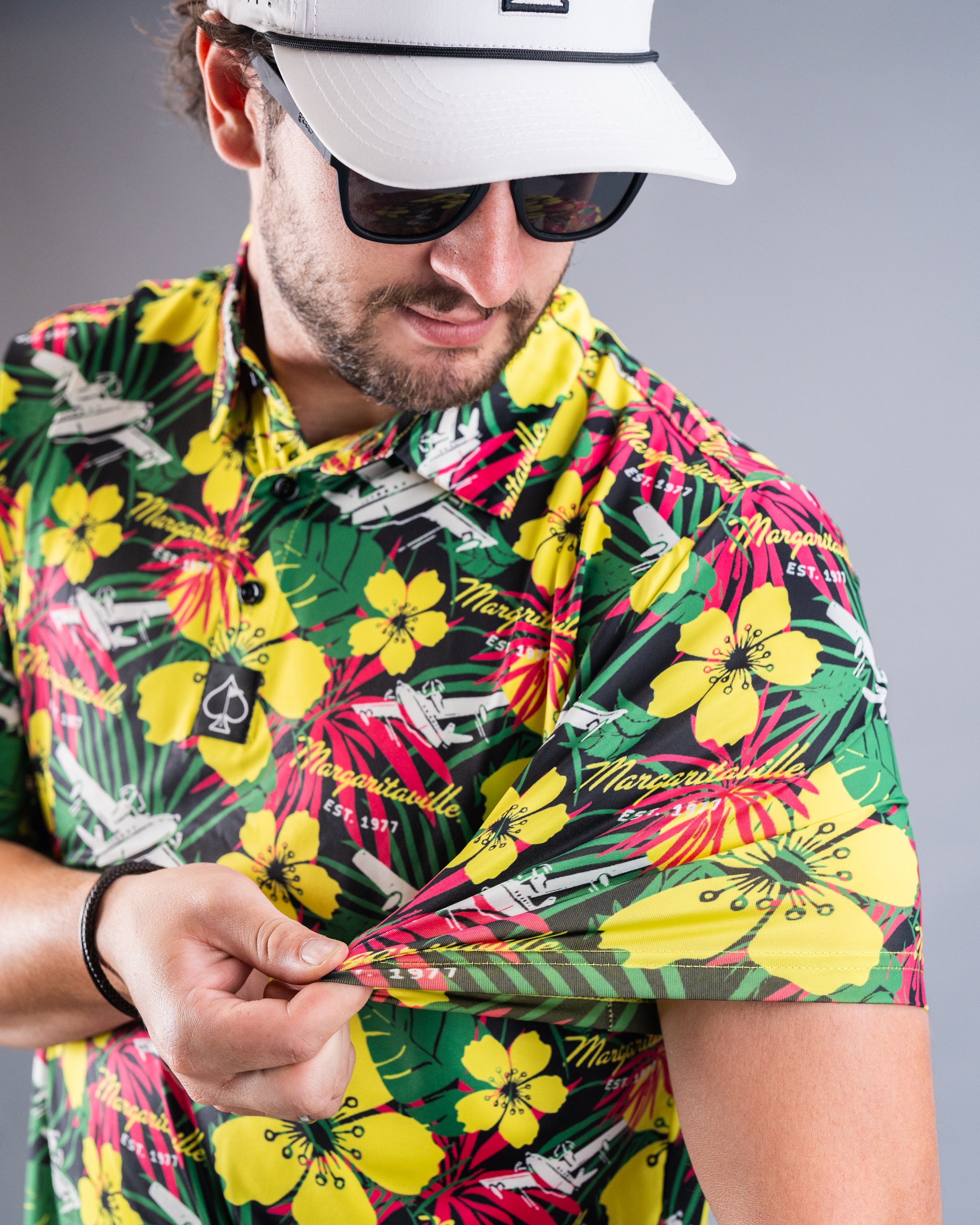Man in tropical floral golf shirt with yellow, pink, and green colors, wearing a white cap and sunglasses, showcasing bold...