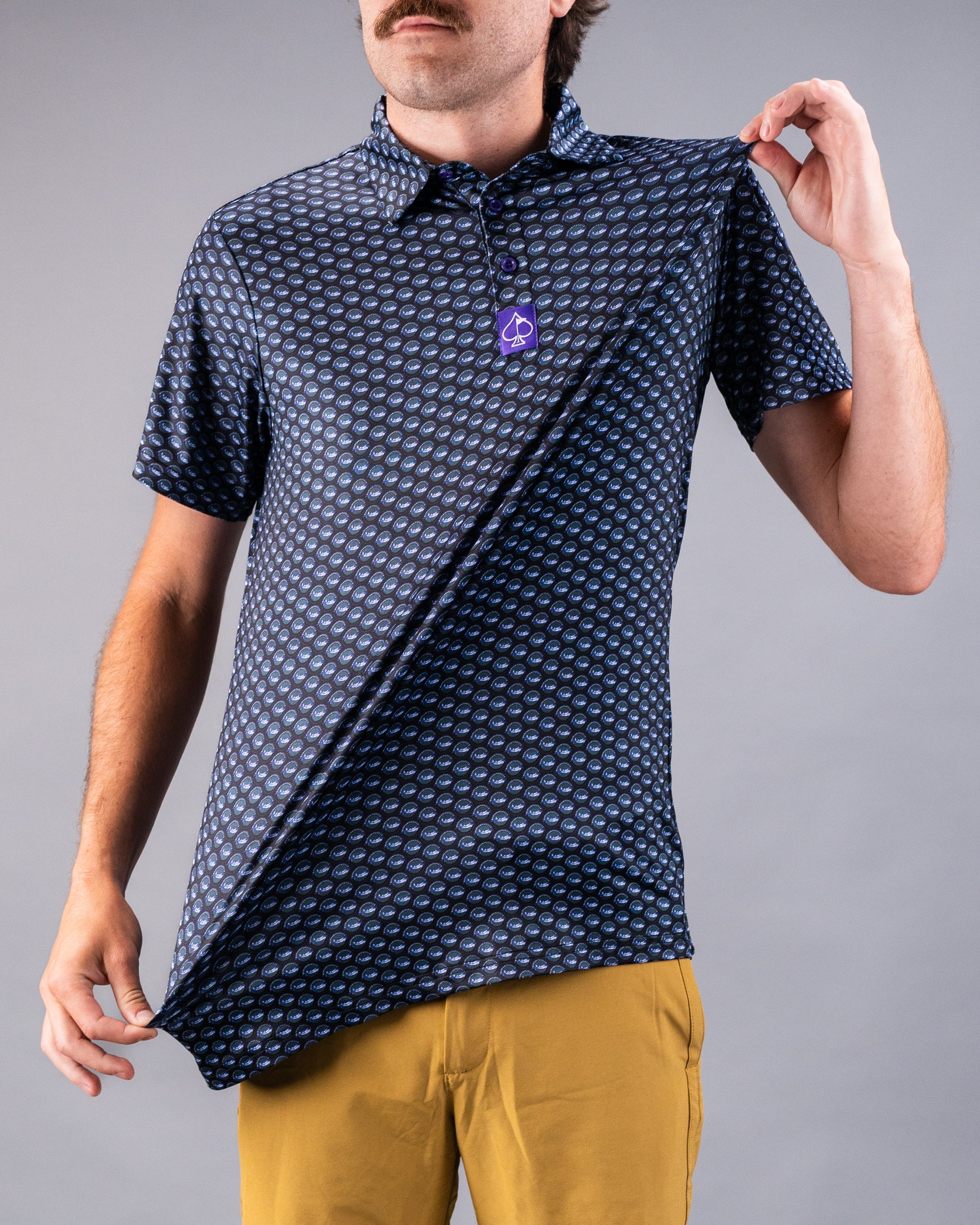 Men's navy golf shirt with a blue geometric pattern, short sleeves, and a purple logo tag, paired with mustard yellow pants.