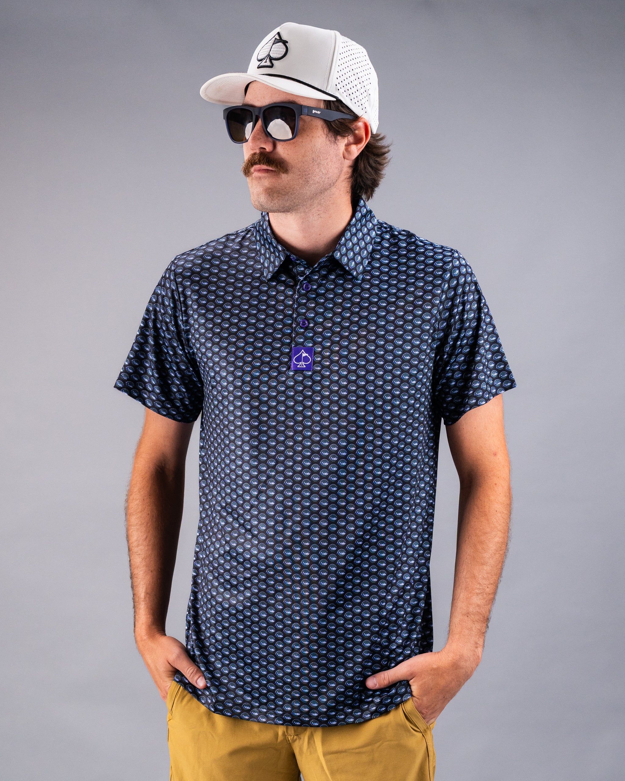 Man wearing a bold navy golf shirt with circular pattern, yellow shorts, black sunglasses, and a white cap with a black logo.