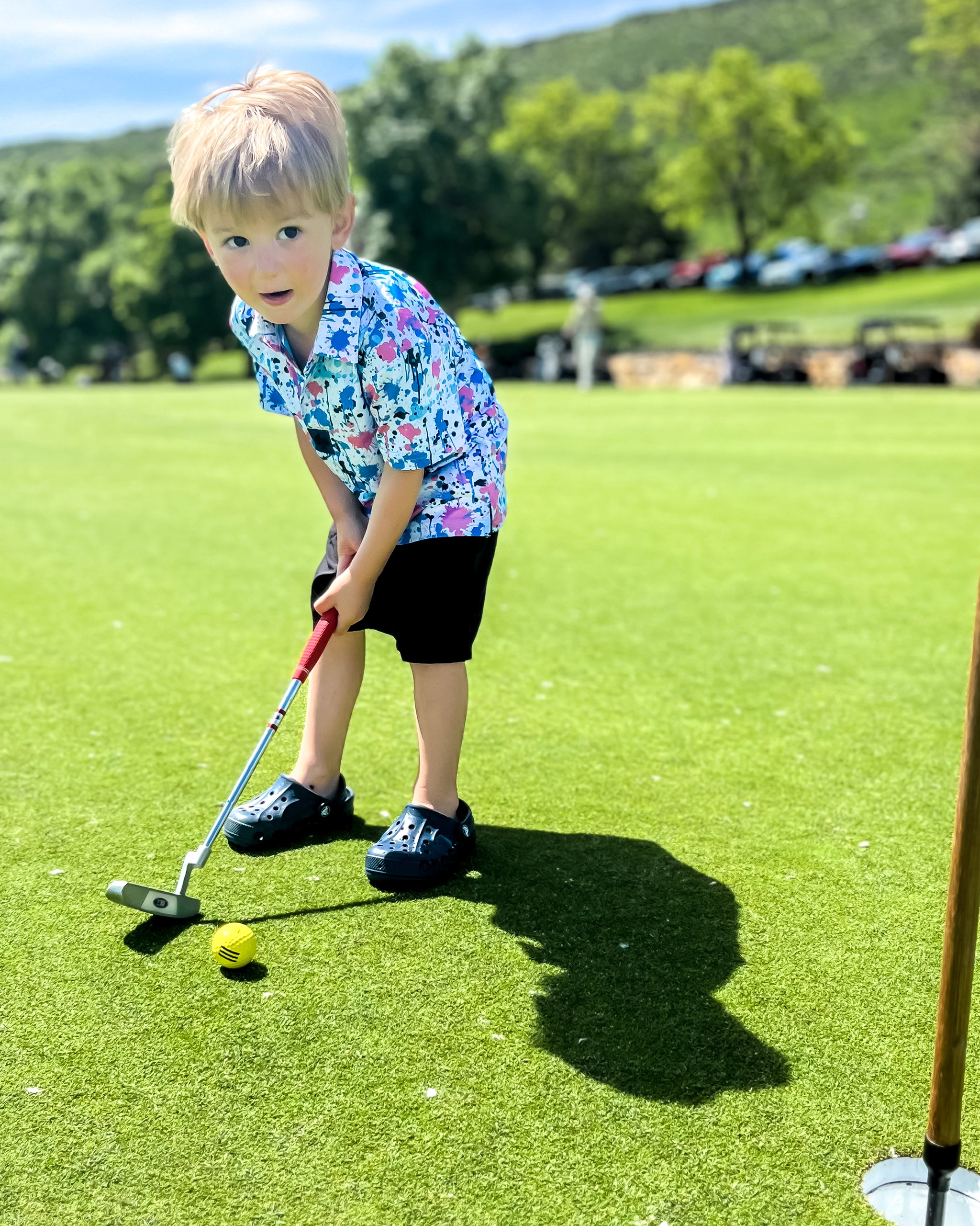 A young boy in a colorful cotton candy drip polo shirt putts a golf ball on a lush green course.