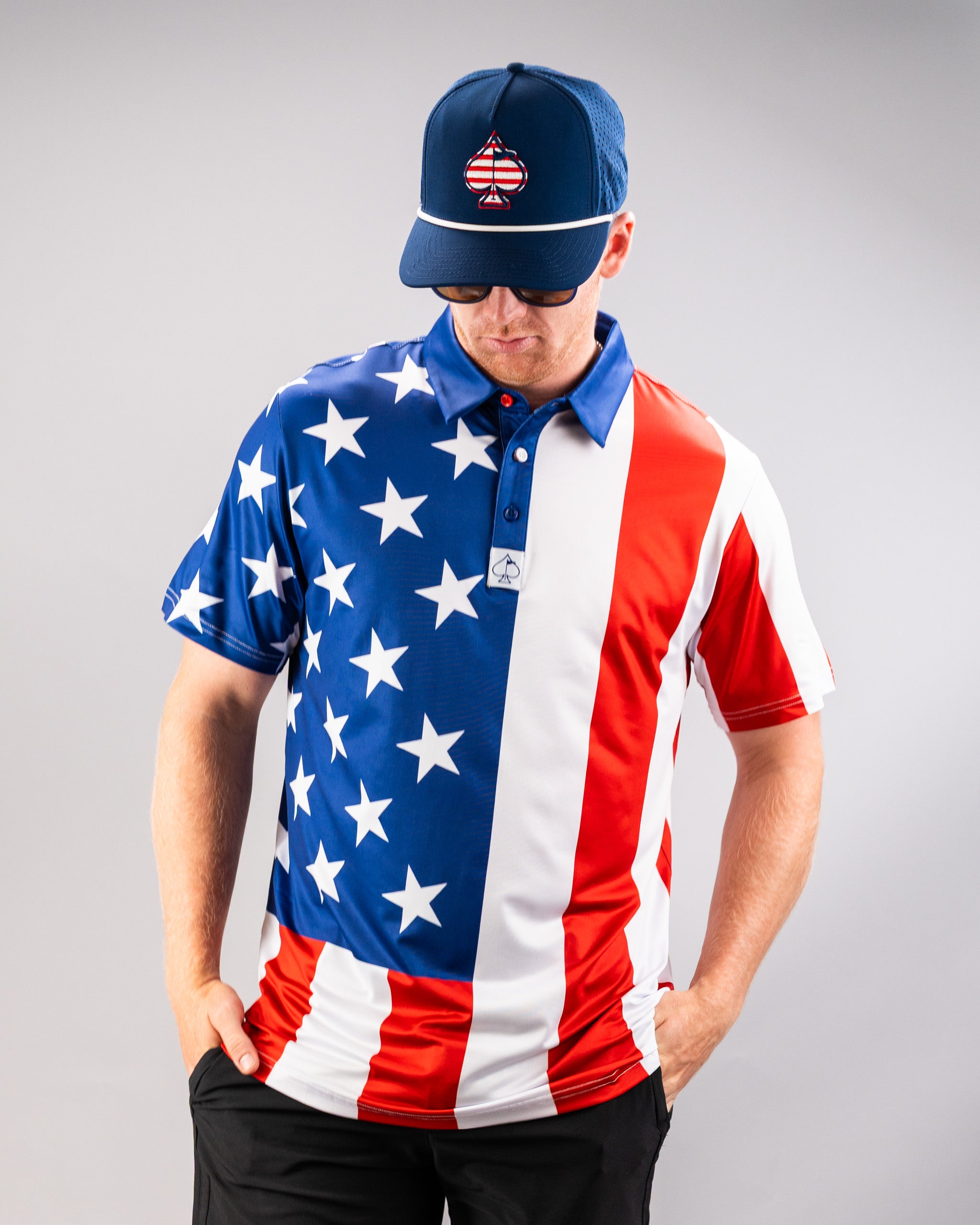 A man wearing a patriotic golf shirt with an American flag design, a matching cap with a spade logo, and sunglasses, standing with hands in pockets.