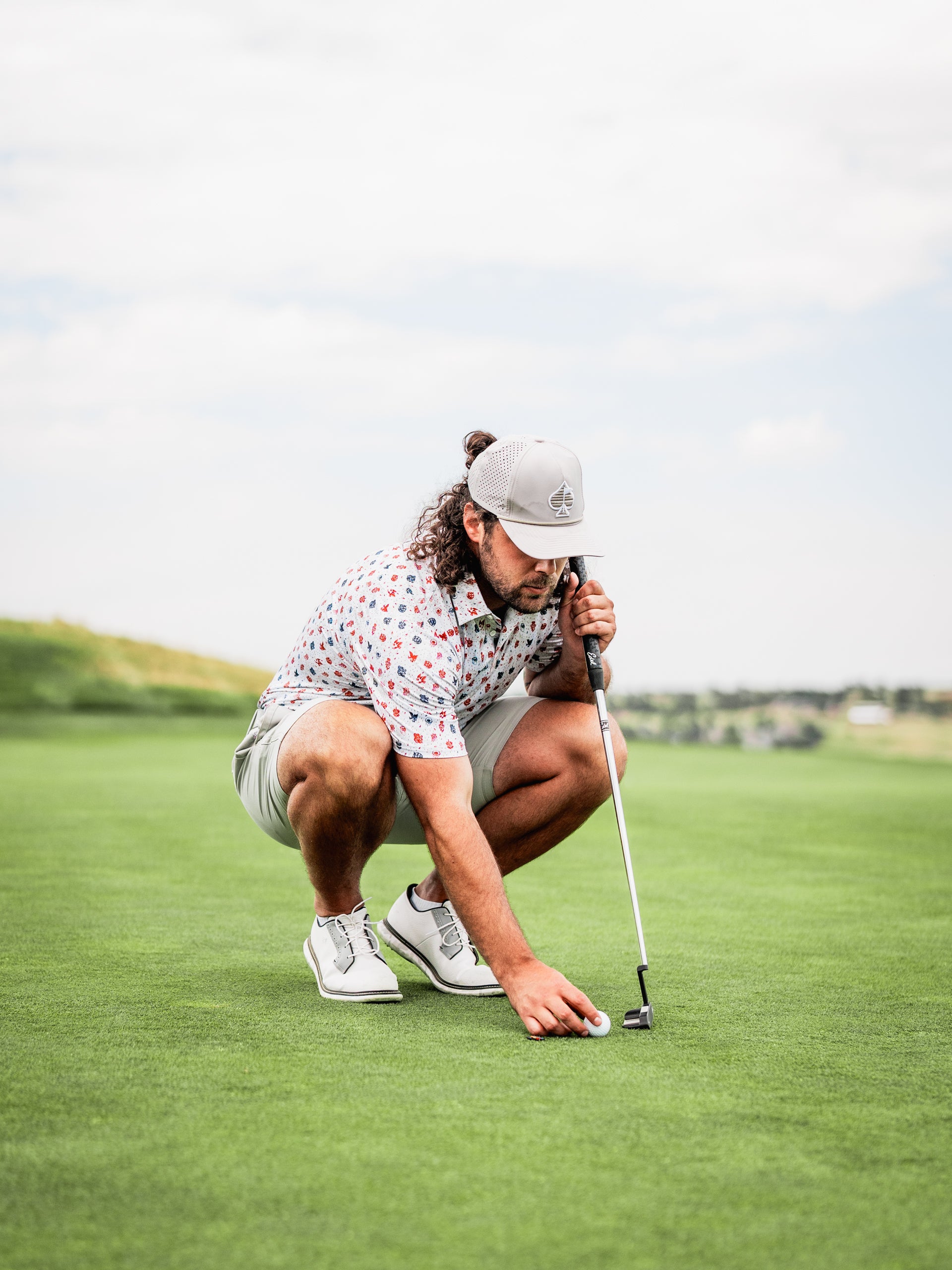 A golfer in a colorful American Traditional shirt kneels on the green, lining up a putt with focused concentration.