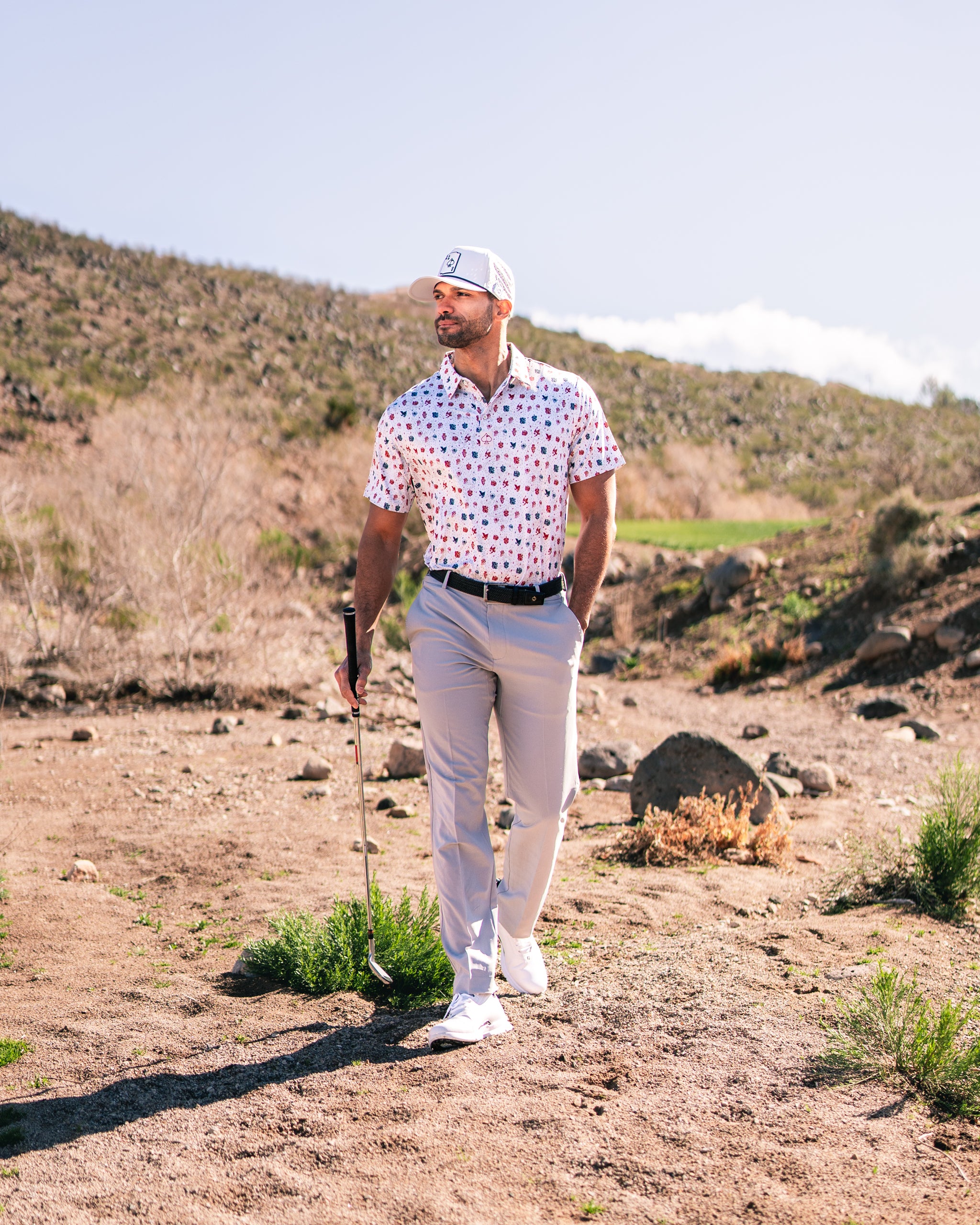 Stylish golfer wearing a patterned polo and light pants, walking on a golf course with a club in hand.