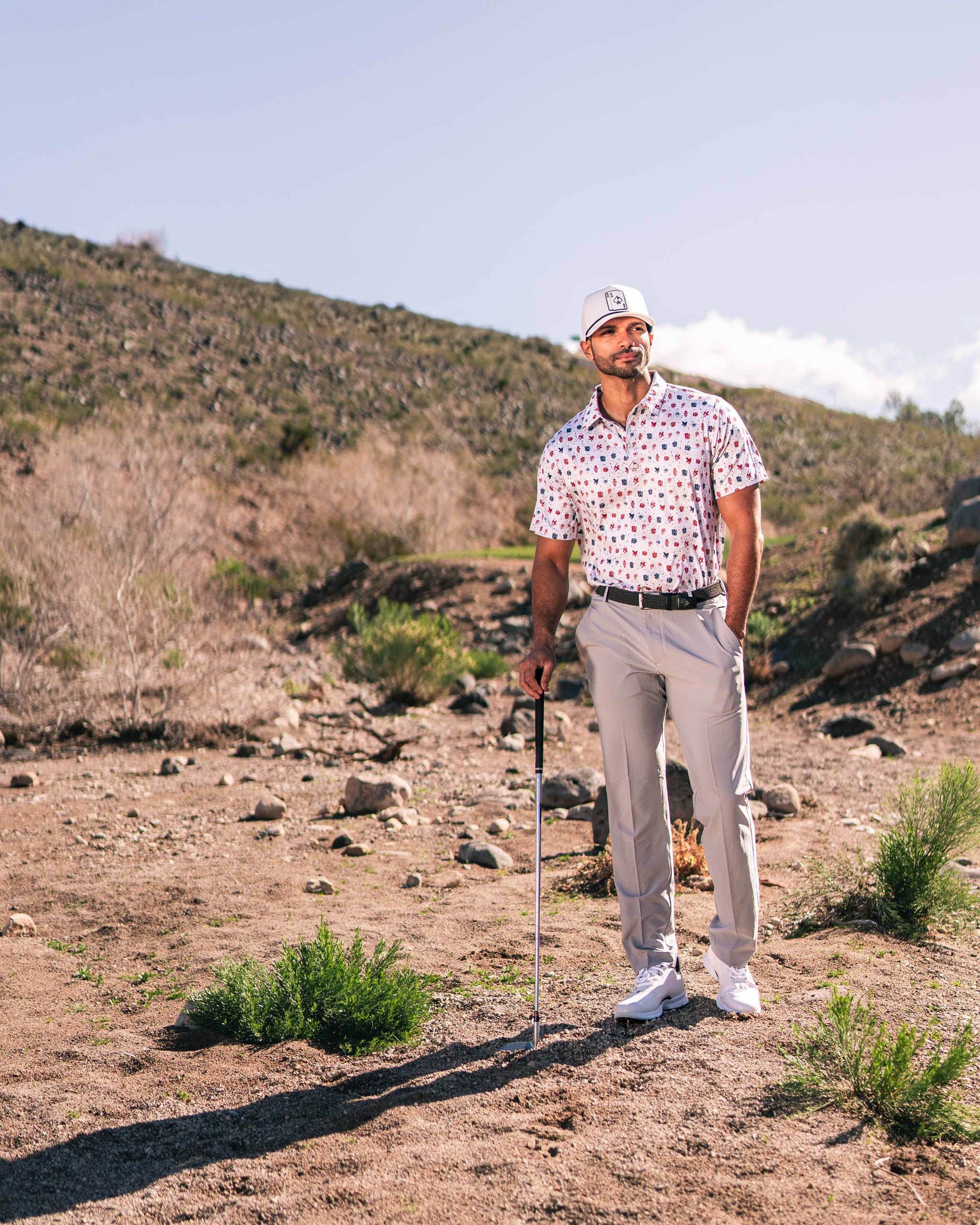 Man wearing a pink and white polka dot golf shirt and light gray pants stands confidently on a rocky golf course.