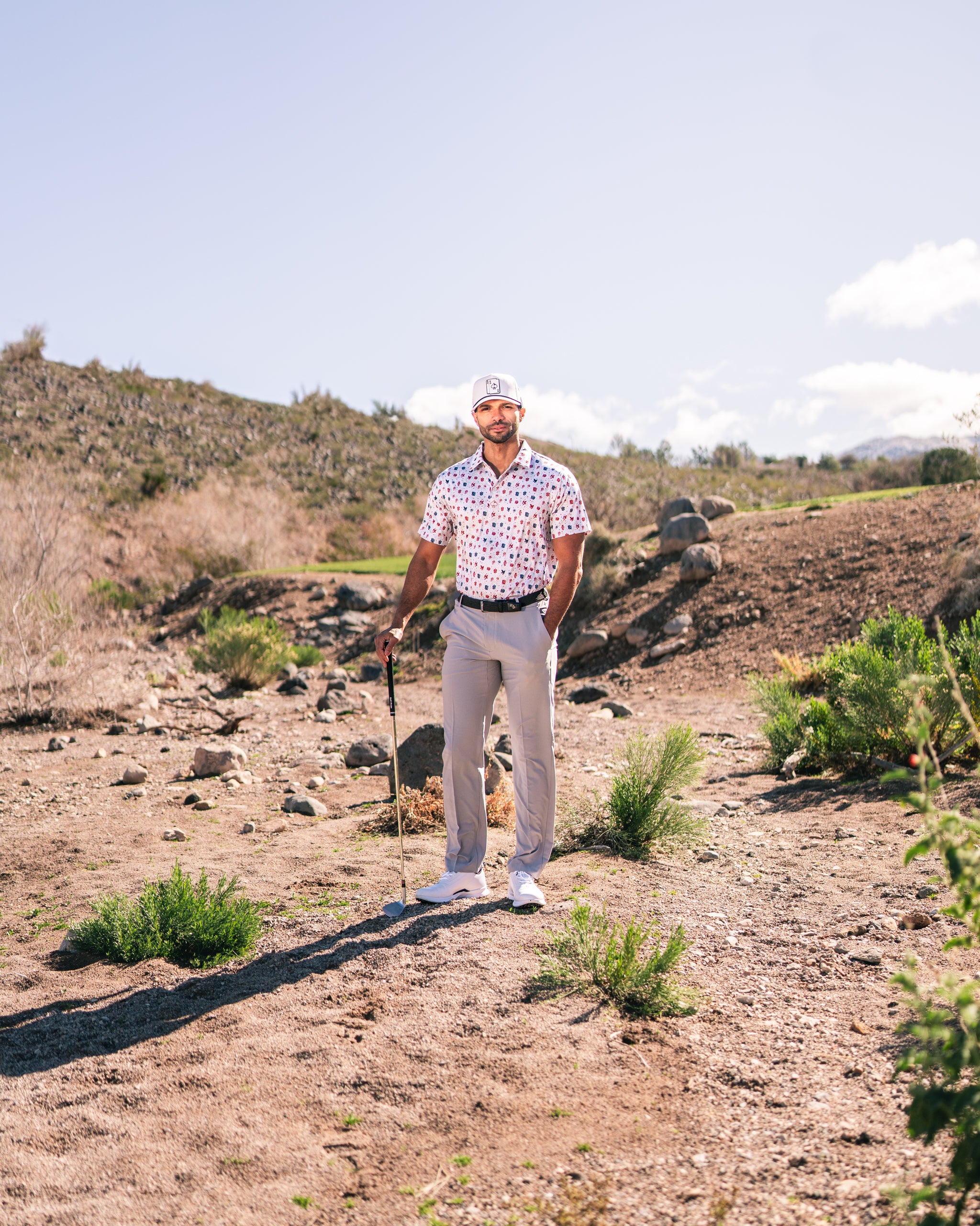 Man wearing a patterned golf shirt and light pants stands confidently on a golf course with a club in hand.