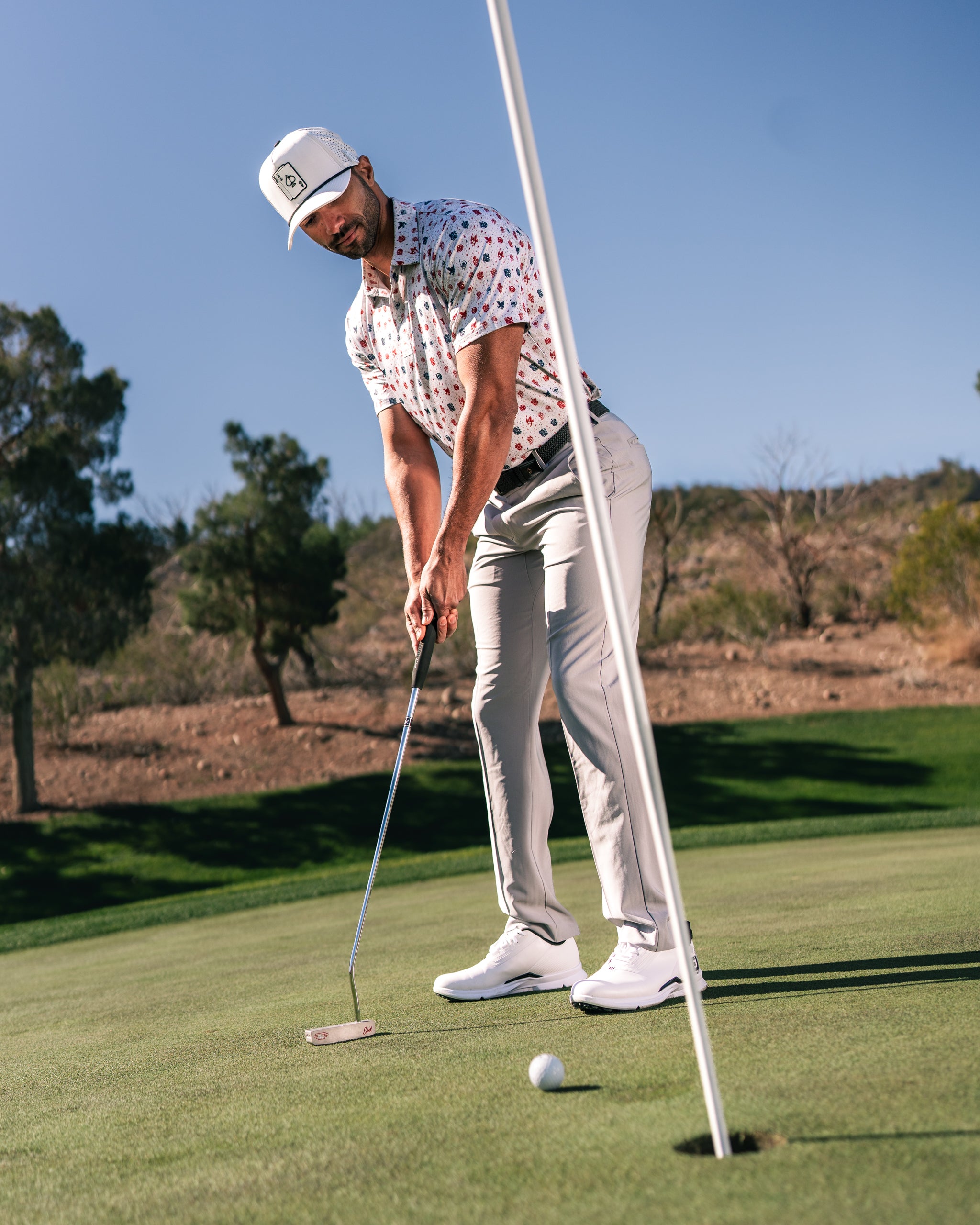 Golfer wearing an American Traditional shirt and cap, preparing to putt on a sunny green course.