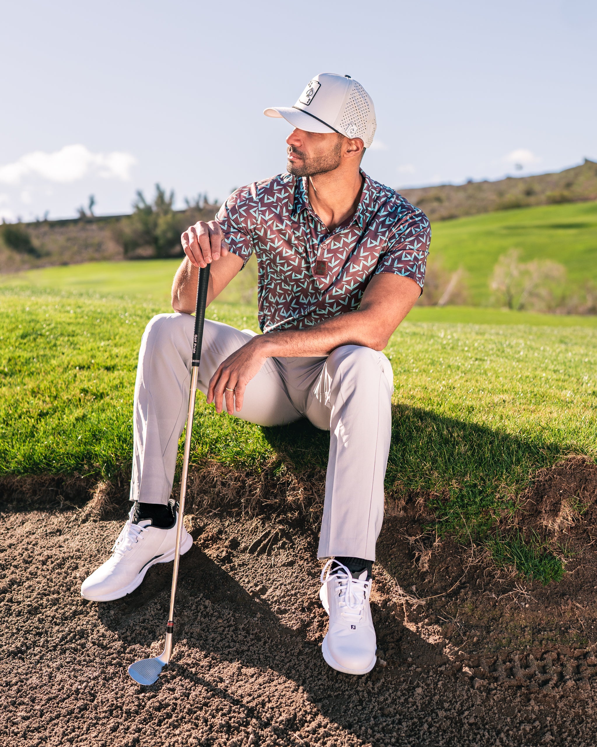Stylish golfer in Cocoa Verde polo shirt and light pants, sitting on a sand trap with a golf club, set against a green course backdrop.