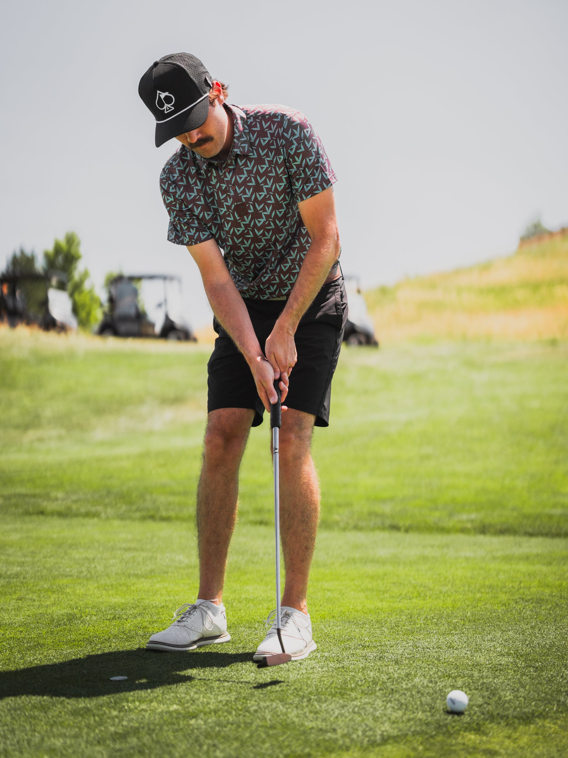 Golfer wearing a patterned shirt and cap, preparing to putt on a sunny course with golf carts in the background.