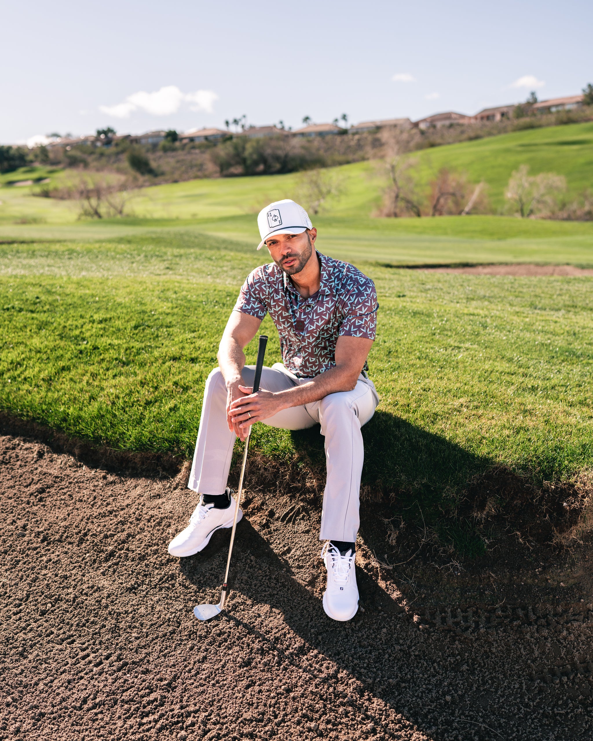 A golfer in a colorful Cocoa Verde shirt sits on the edge of a sand trap, holding a club, with a lush green course in the background.