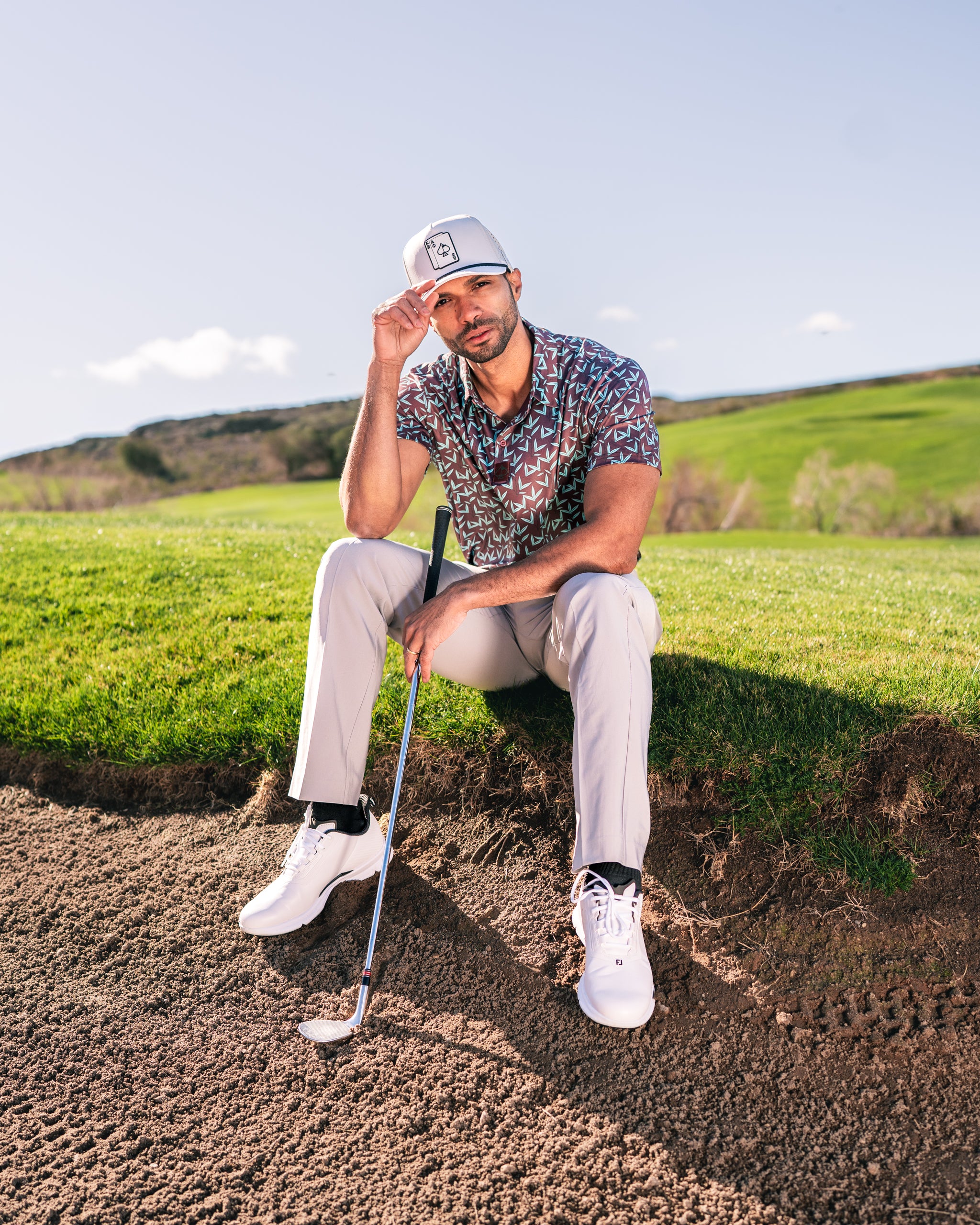 Man sitting on a sand trap wearing a patterned golf shirt and cap, holding a golf club, with a scenic green background.