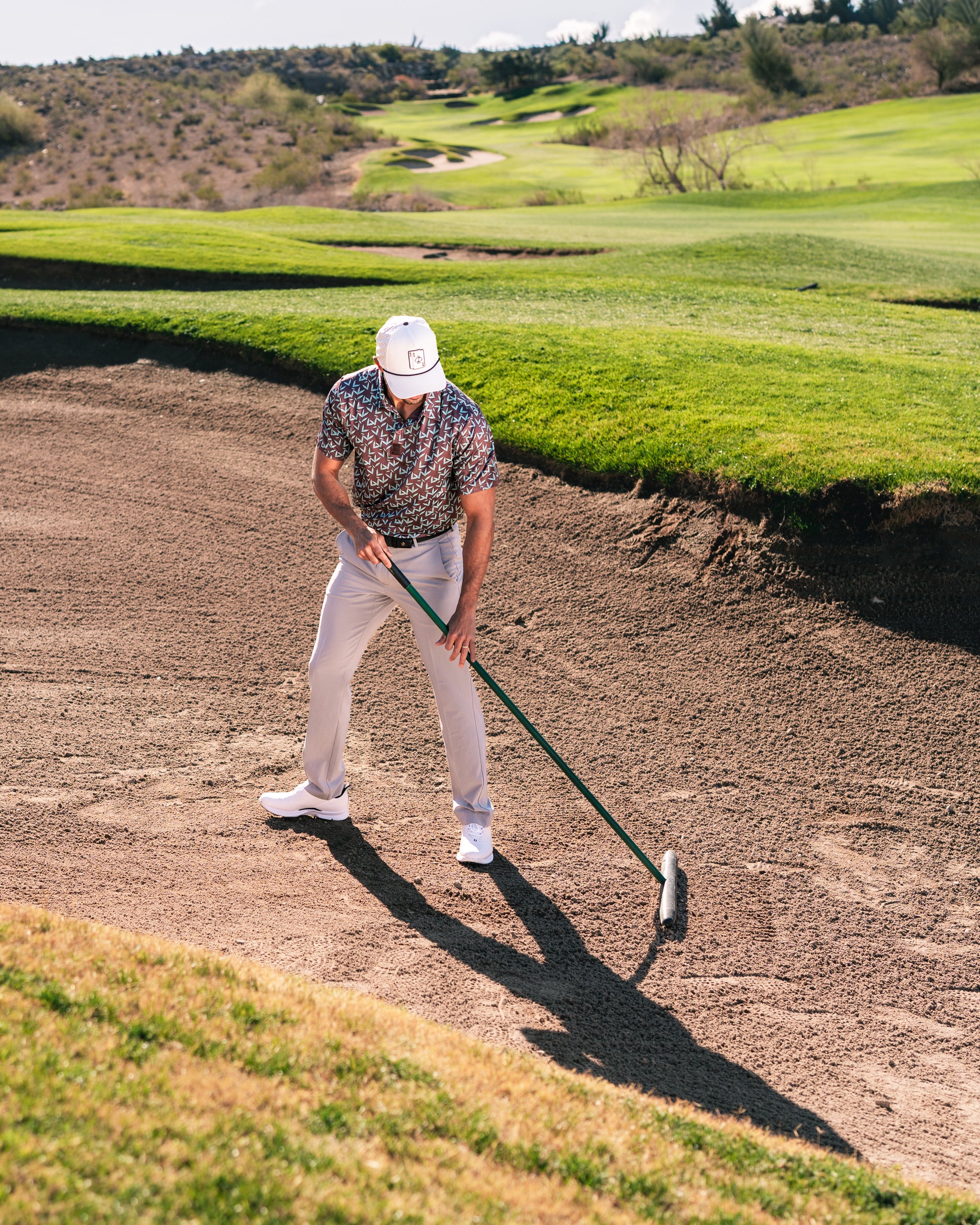 A golfer in a patterned shirt and white hat rakes a sand trap on a sunny golf course, showcasing Cocoa Verde apparel.