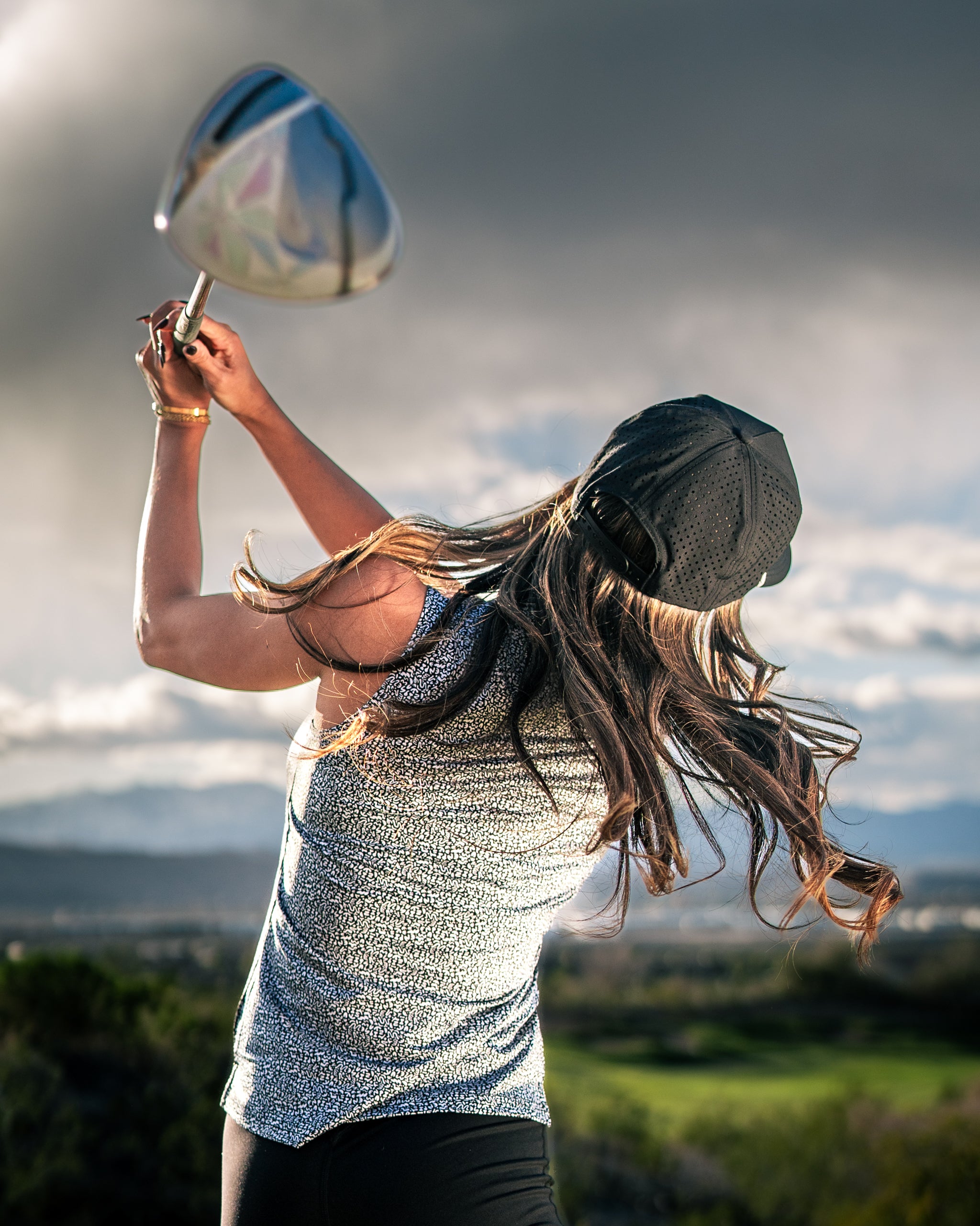 A woman swings a golf club, wearing a stylish coral top and a cap, with a scenic landscape in the background.