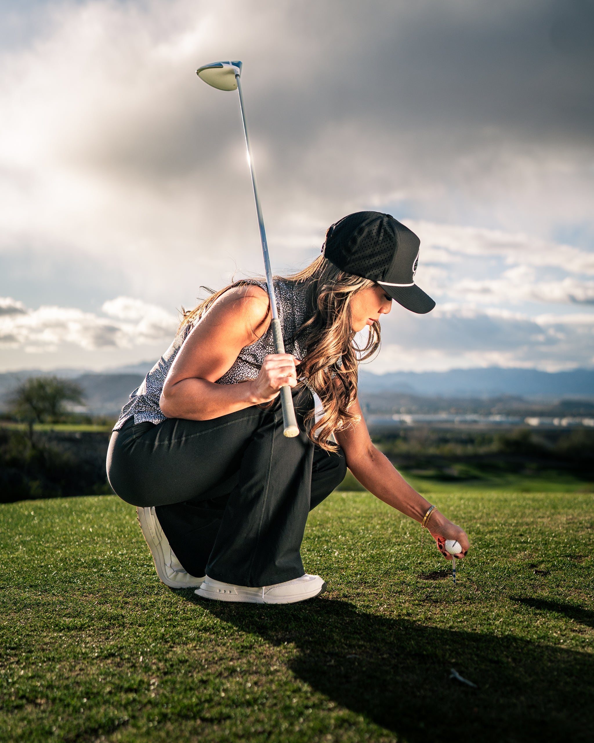 A woman in a stylish golf outfit crouches on the green, preparing to tee off with a golf club in hand.