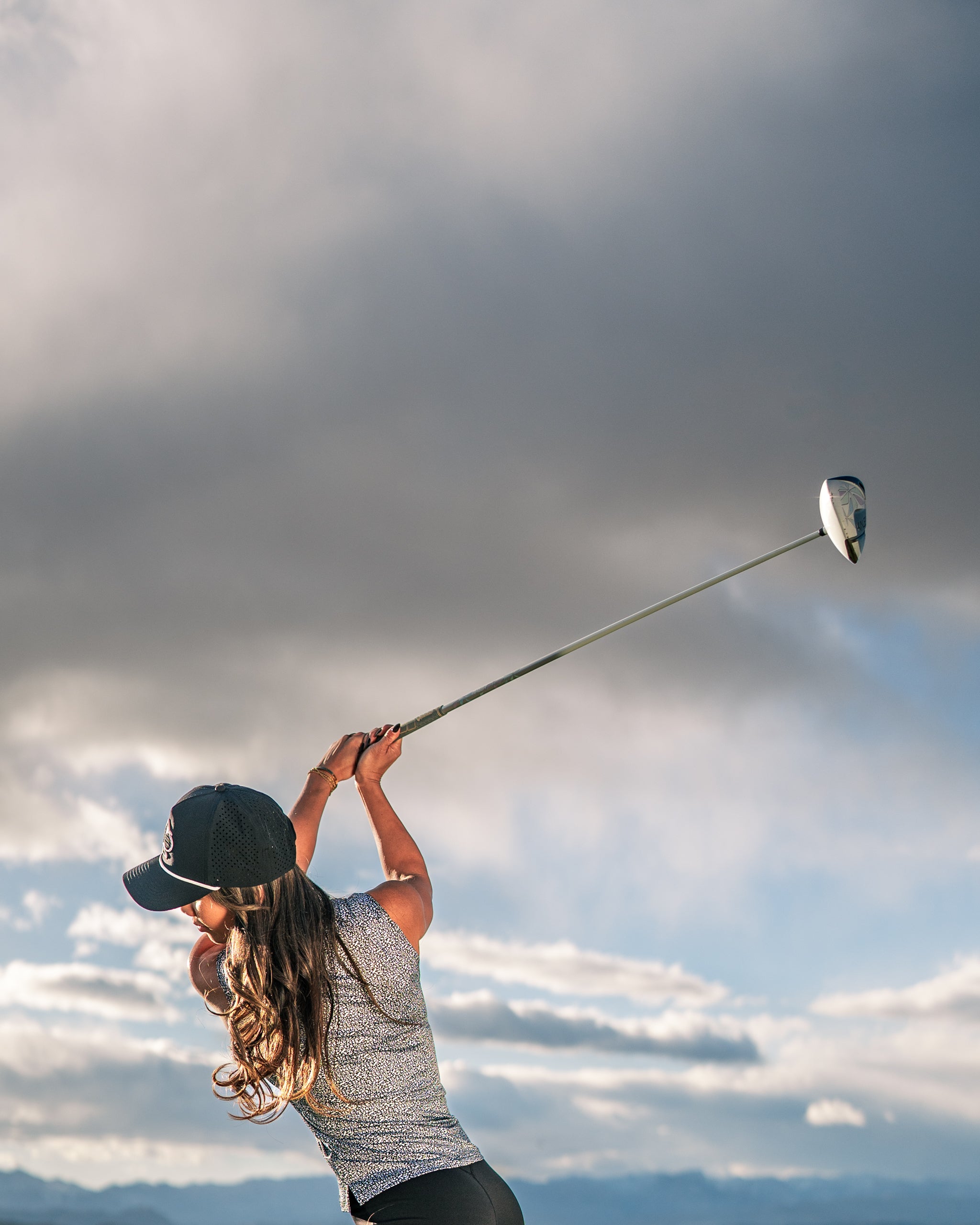 Woman in a stylish coral golf shirt swings a driver against a dramatic sky, showcasing bold golf apparel.