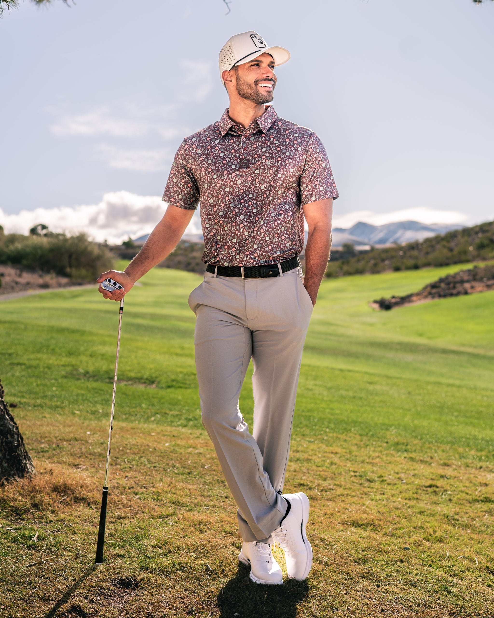 Man in a floral golf shirt and light pants stands on a golf course, smiling with a club in hand and mountains in the background.