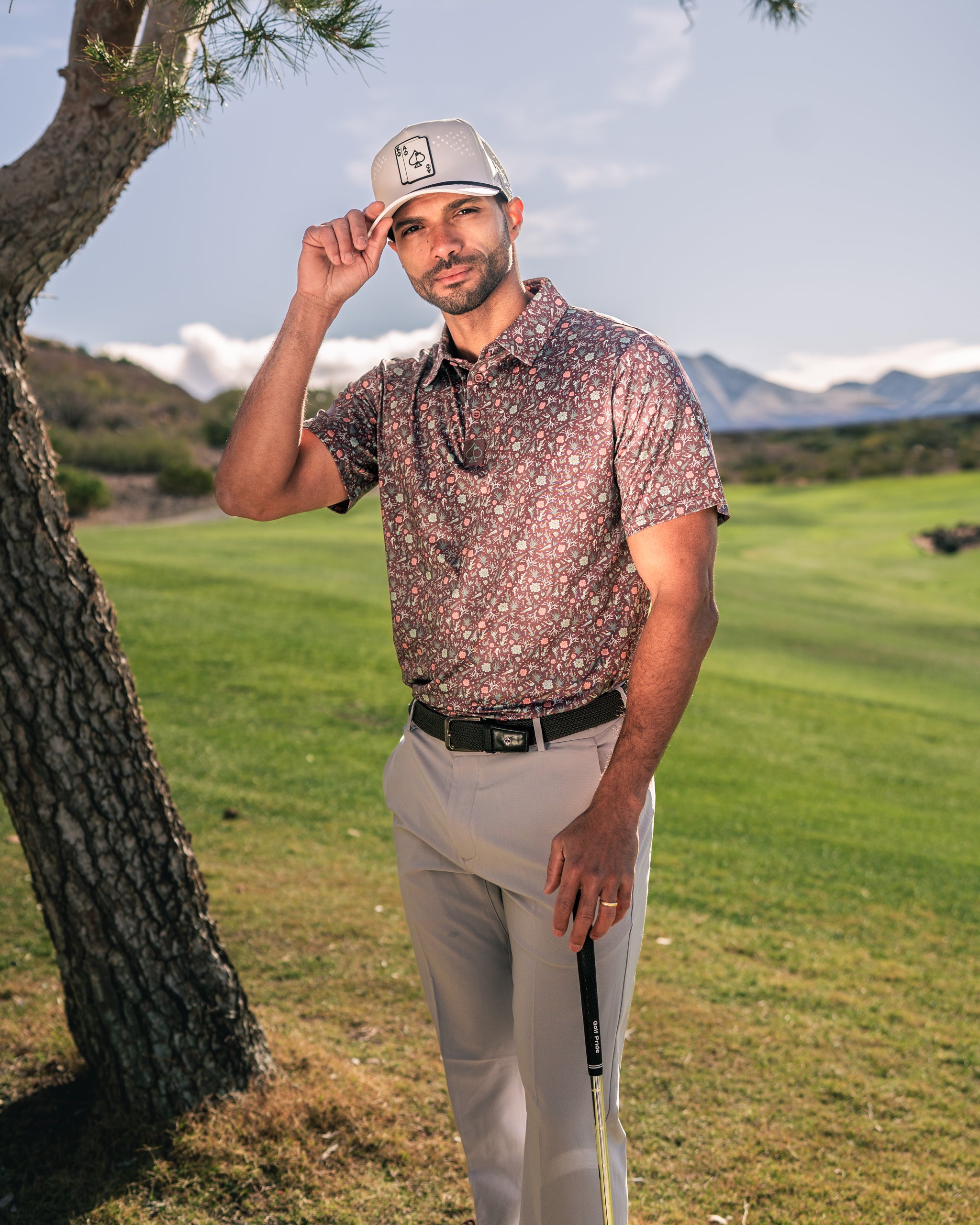 A golfer wearing a floral patterned shirt and a cap poses confidently on a golf course with mountains in the background.