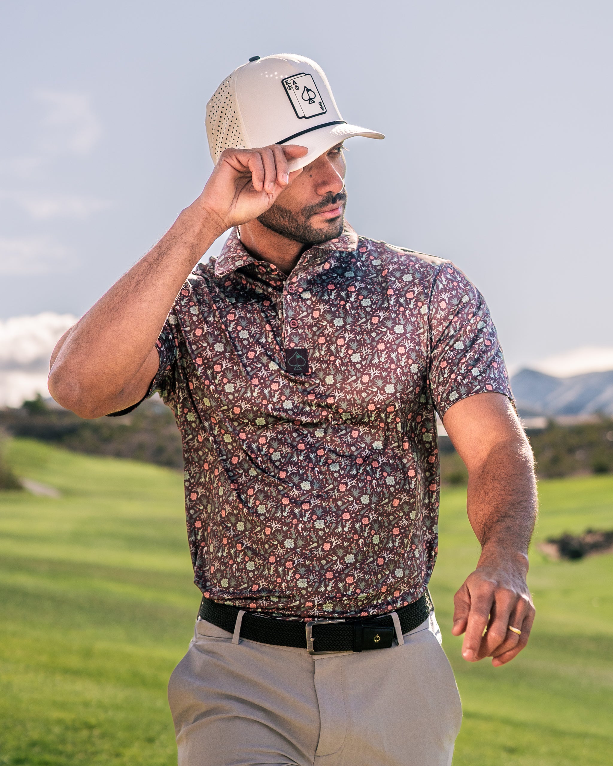 Stylish golfer wearing a Desert Sage patterned polo and a light cap, walking on a lush golf course under a clear sky.