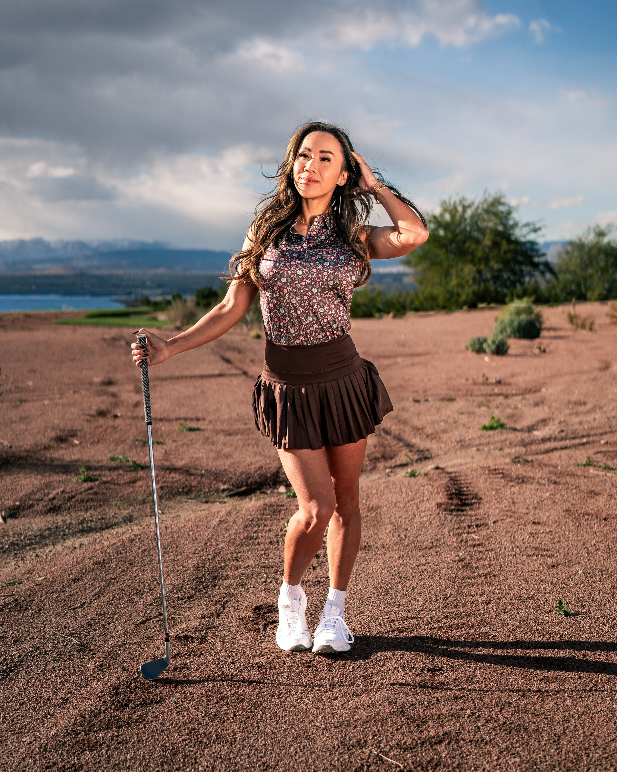 Stylish woman in a floral golf shirt and pleated skirt poses confidently on a desert landscape, holding a golf club.