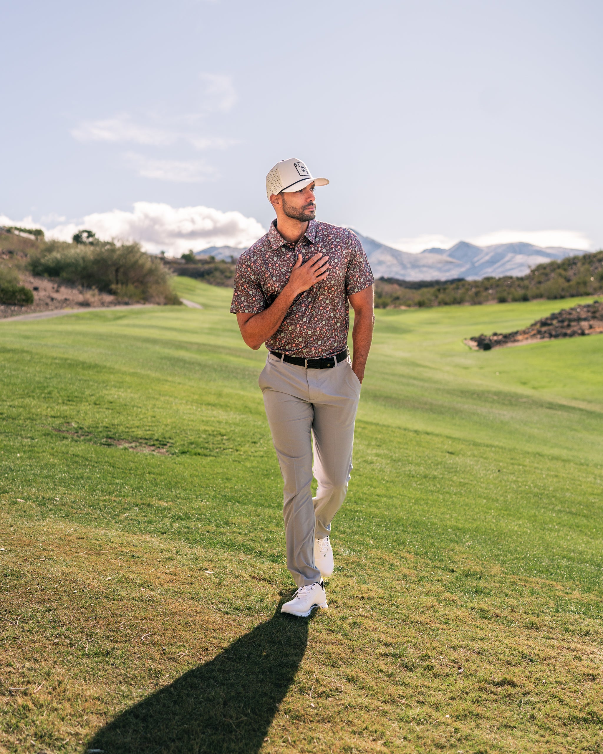 A golfer in a floral Desert Sage shirt and beige pants walks confidently on a lush green golf course under a clear sky.