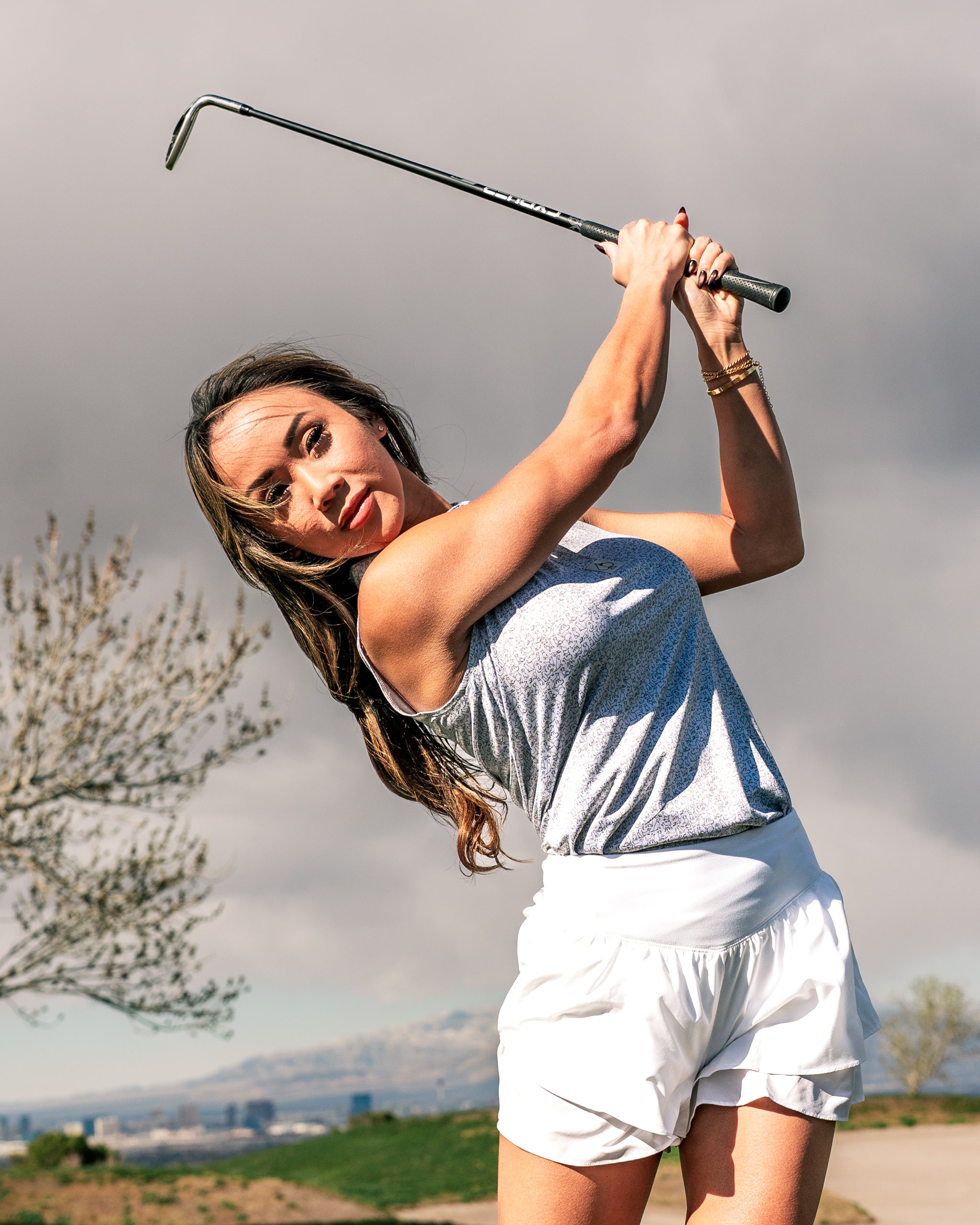 A woman swings a golf club on a course, wearing a stylish light gray top and white shorts, showcasing bold golf fashion.