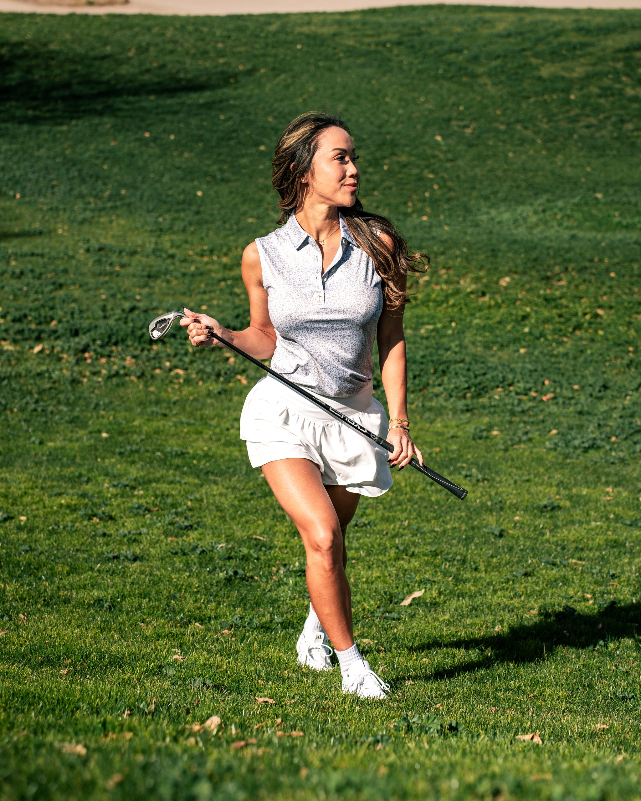 Woman in a stylish golf polo and skirt walks confidently on the course, holding a golf club against a green backdrop.