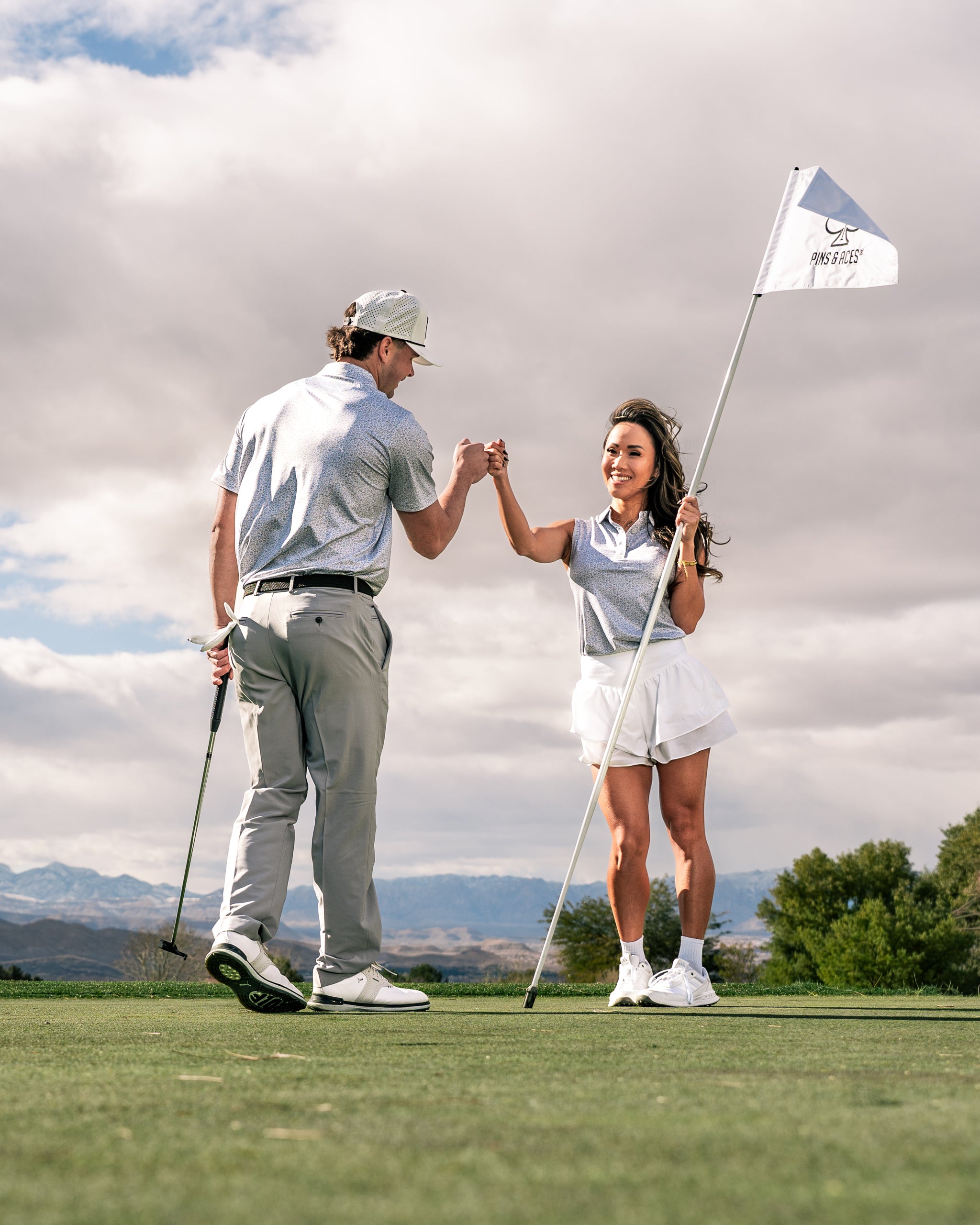 A woman in a stylish golf outfit gives a fist bump to a man on the course, with a golf flag in the background.