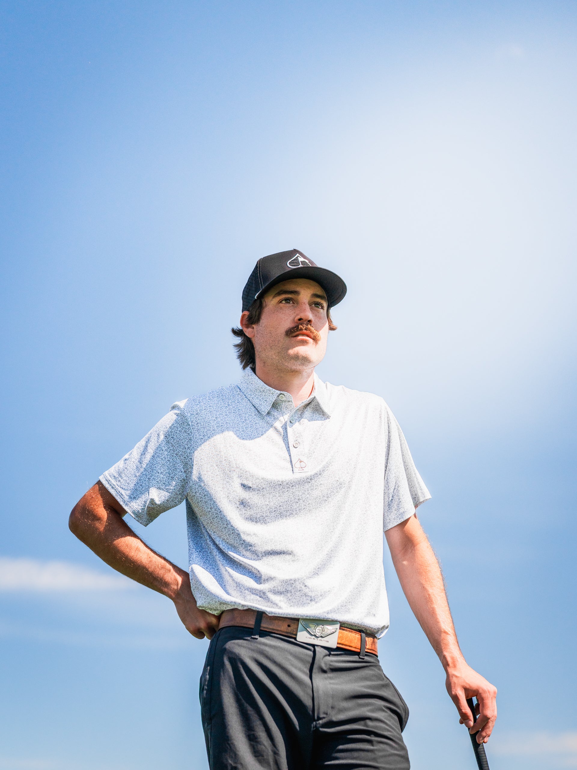 A golfer wearing a light-patterned polo shirt and cap stands confidently on the course, holding a golf club.