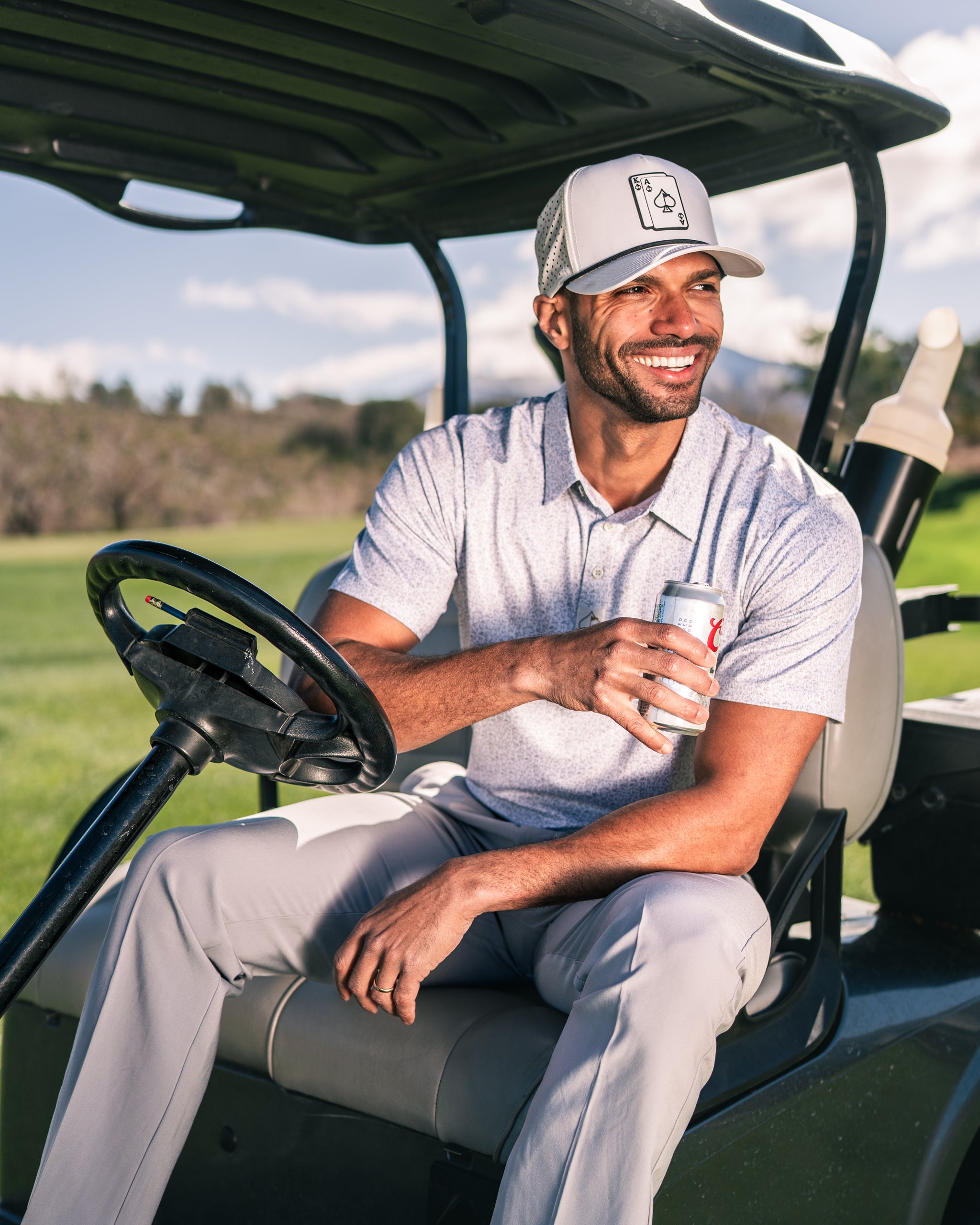 Man enjoying a drink while seated in a golf cart, wearing a stylish polo shirt and cap on a sunny golf course.