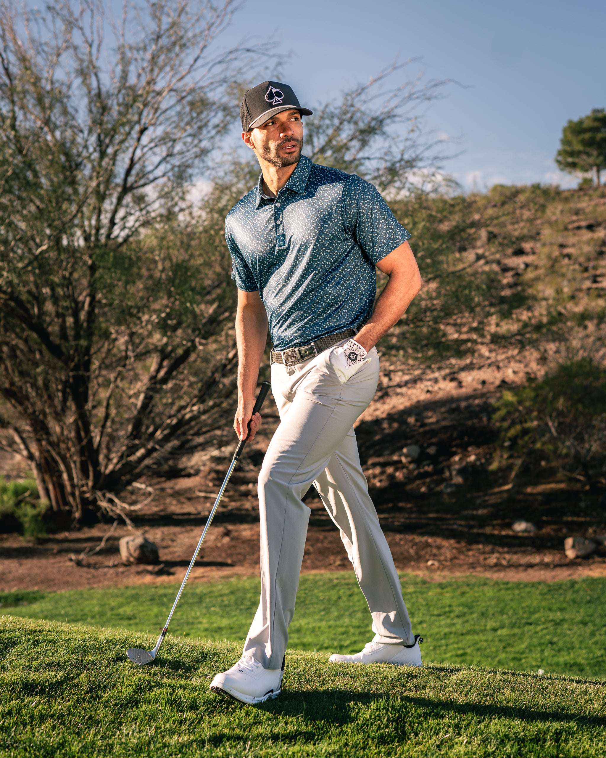 A golfer in a stylish blue patterned polo and light beige pants walks confidently on the course, holding a golf club.