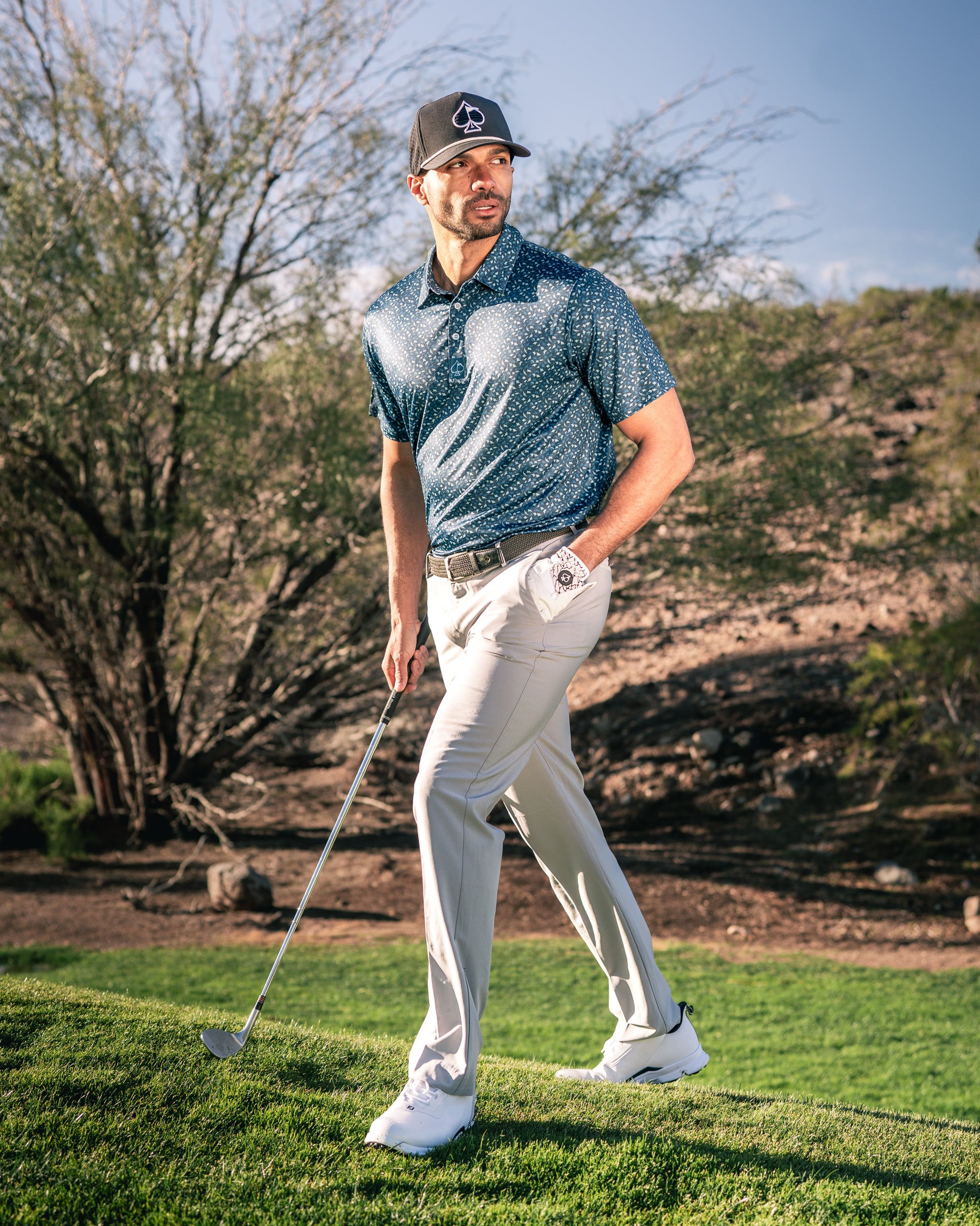 Stylish golfer wearing a patterned polo and light pants, walking confidently on a green course with a golf club.