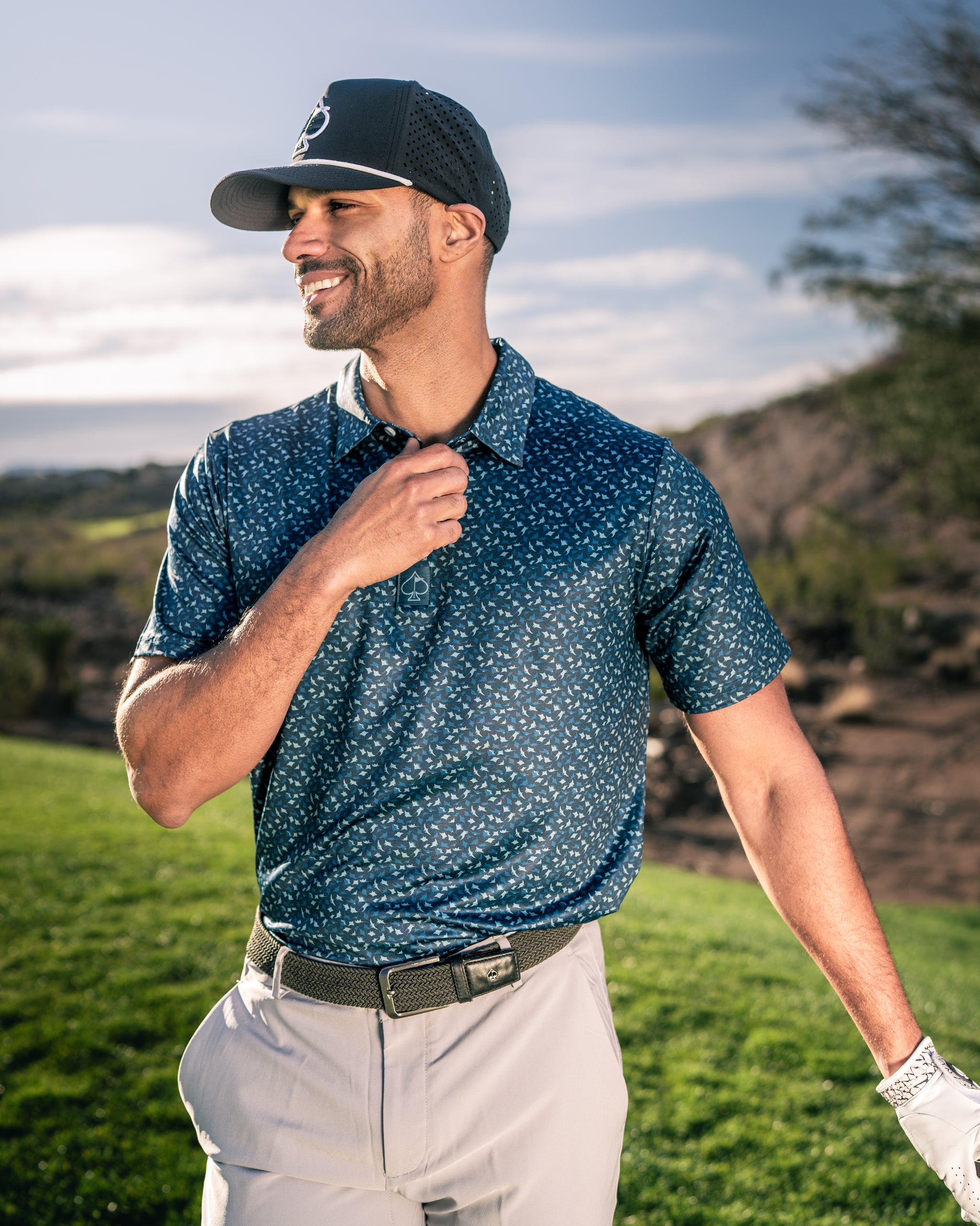 A golfer wearing a navy blue patterned polo shirt and a cap smiles confidently on the course, showcasing bold golf style.