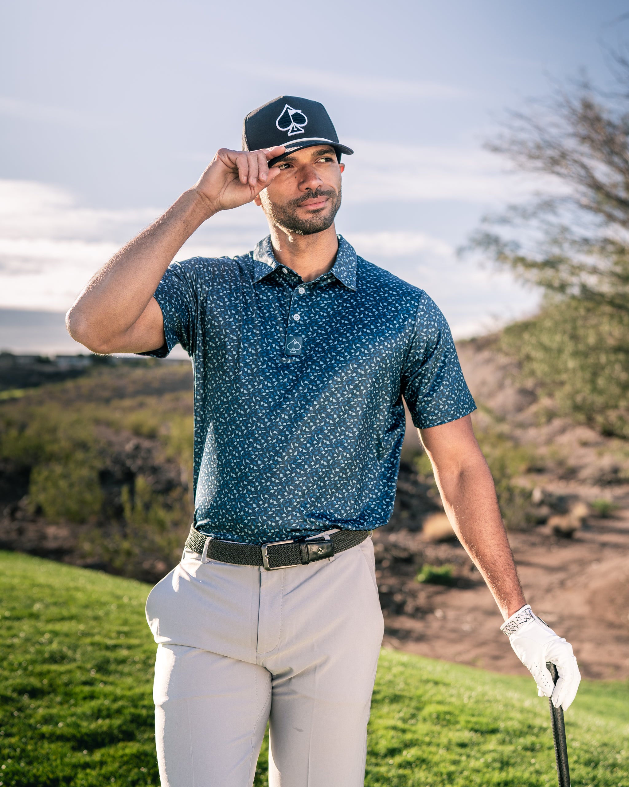 Man wearing a navy blue golf polo with a subtle pattern, adjusting his cap while holding a golf club on a course.