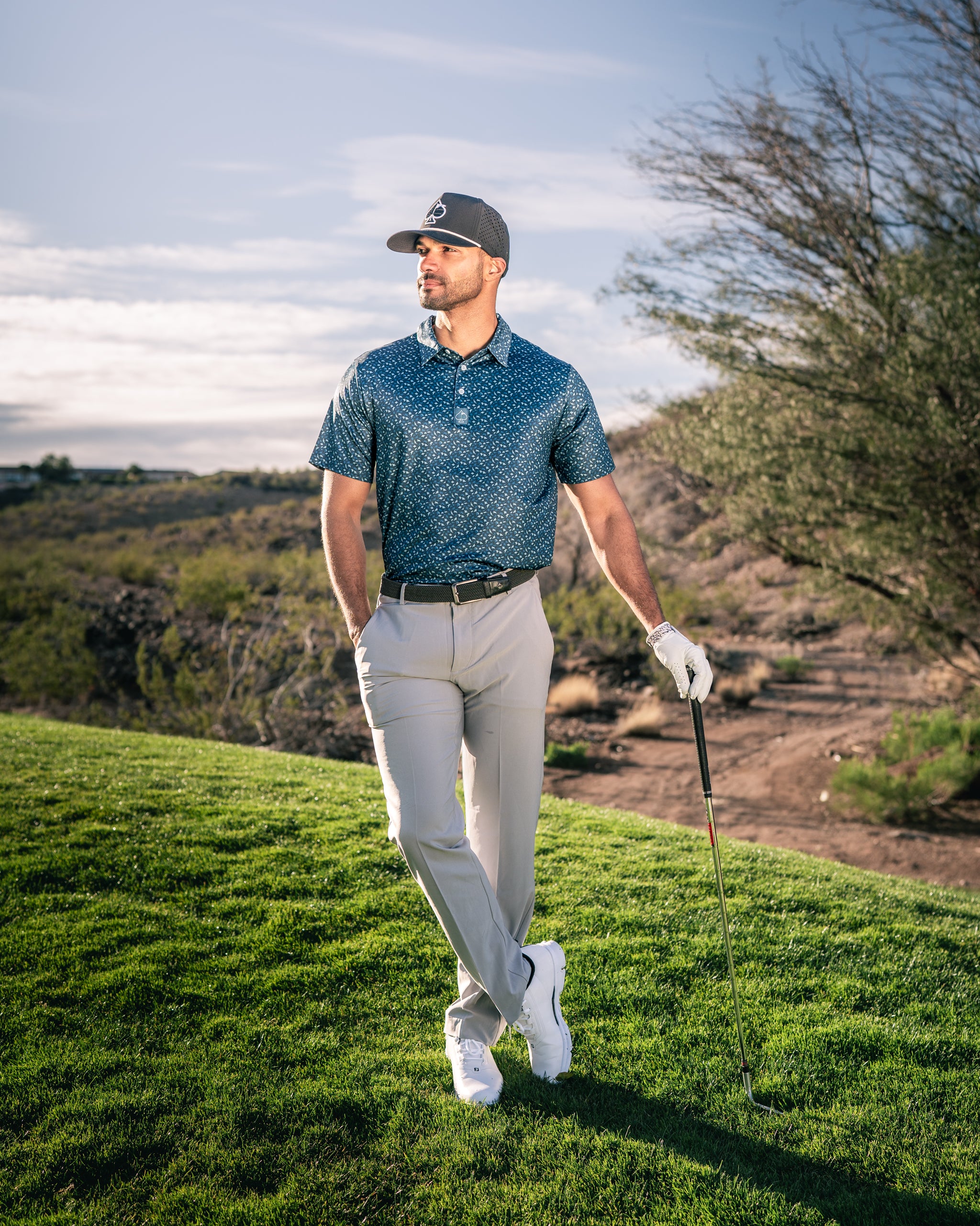 Man in a stylish Manta Ray golf polo and light pants stands on a golf course, holding a club, with a scenic background.