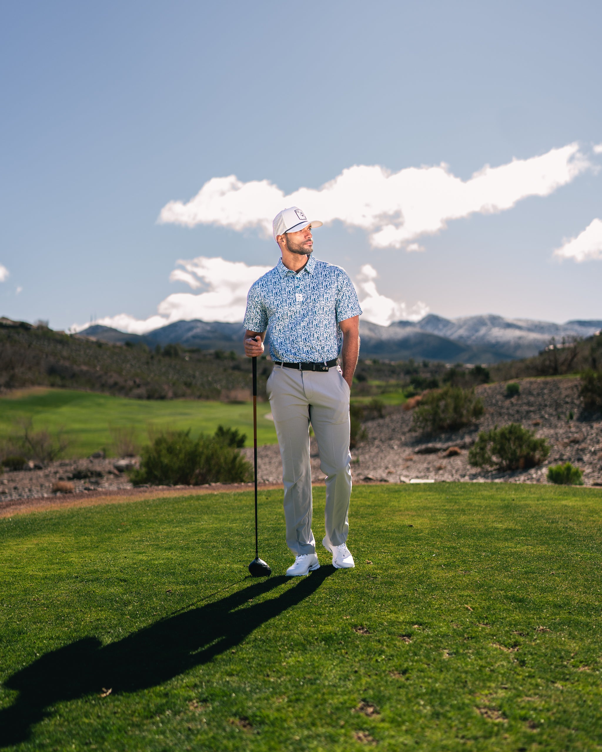 Man in a patterned golf shirt and white cap stands on a green golf course, holding a driver with mountains in the background.