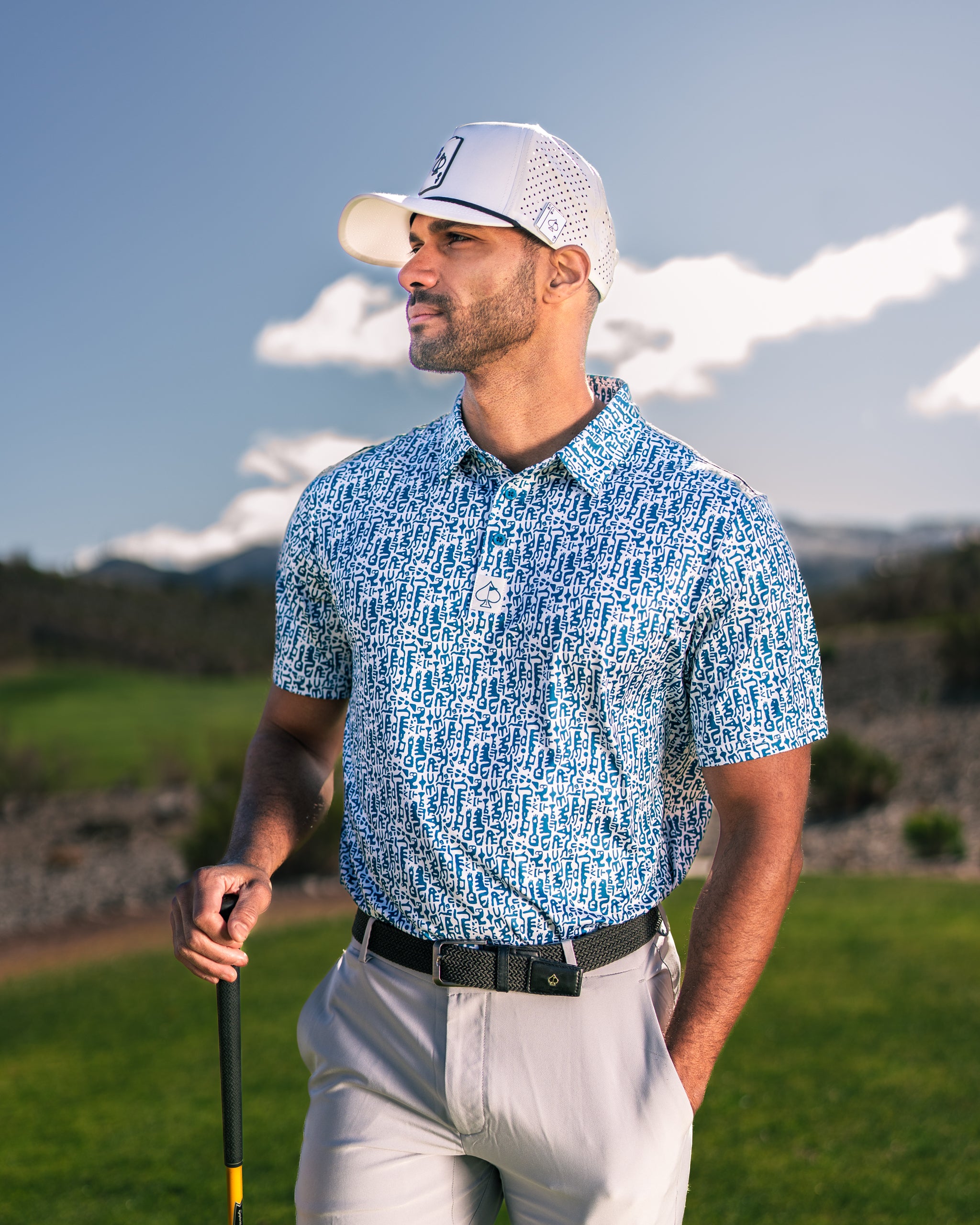 Man wearing a blue and white patterned golf shirt on a sunny course, holding a golf club and looking confidently into the distance.