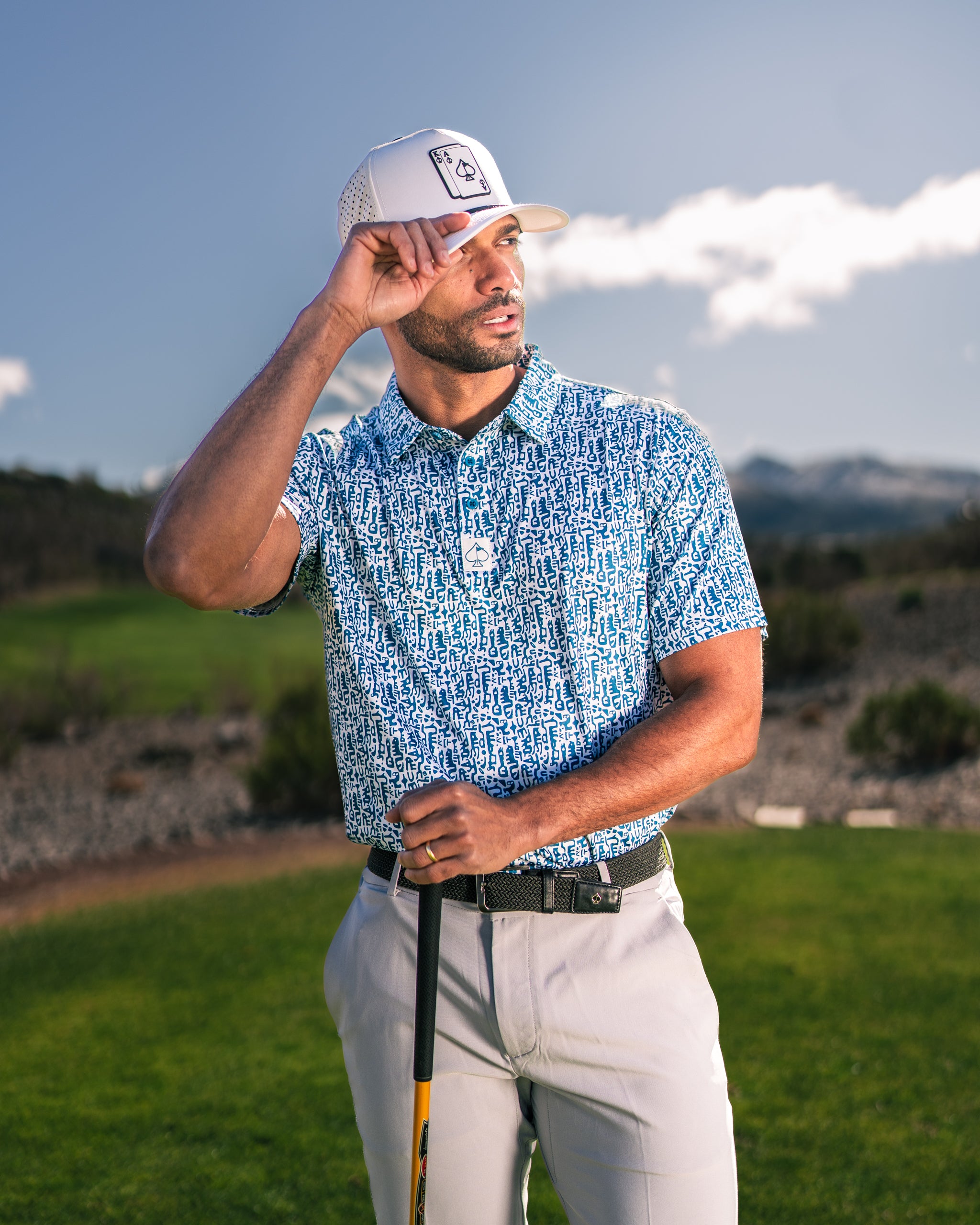 Man in a blue patterned golf shirt and white cap stands on a golf course, holding a club and shielding his eyes from the sun.