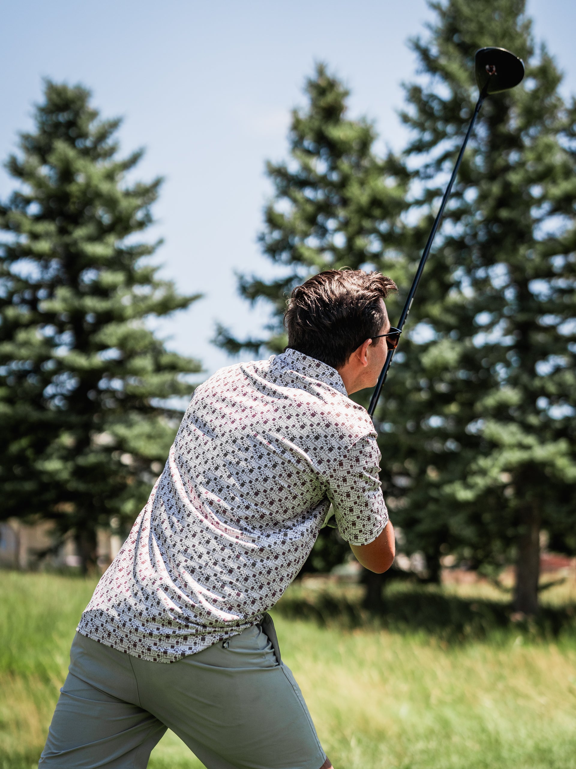 A golfer swings a driver while wearing a stylish retro-patterned shirt and shorts on a sunny course.