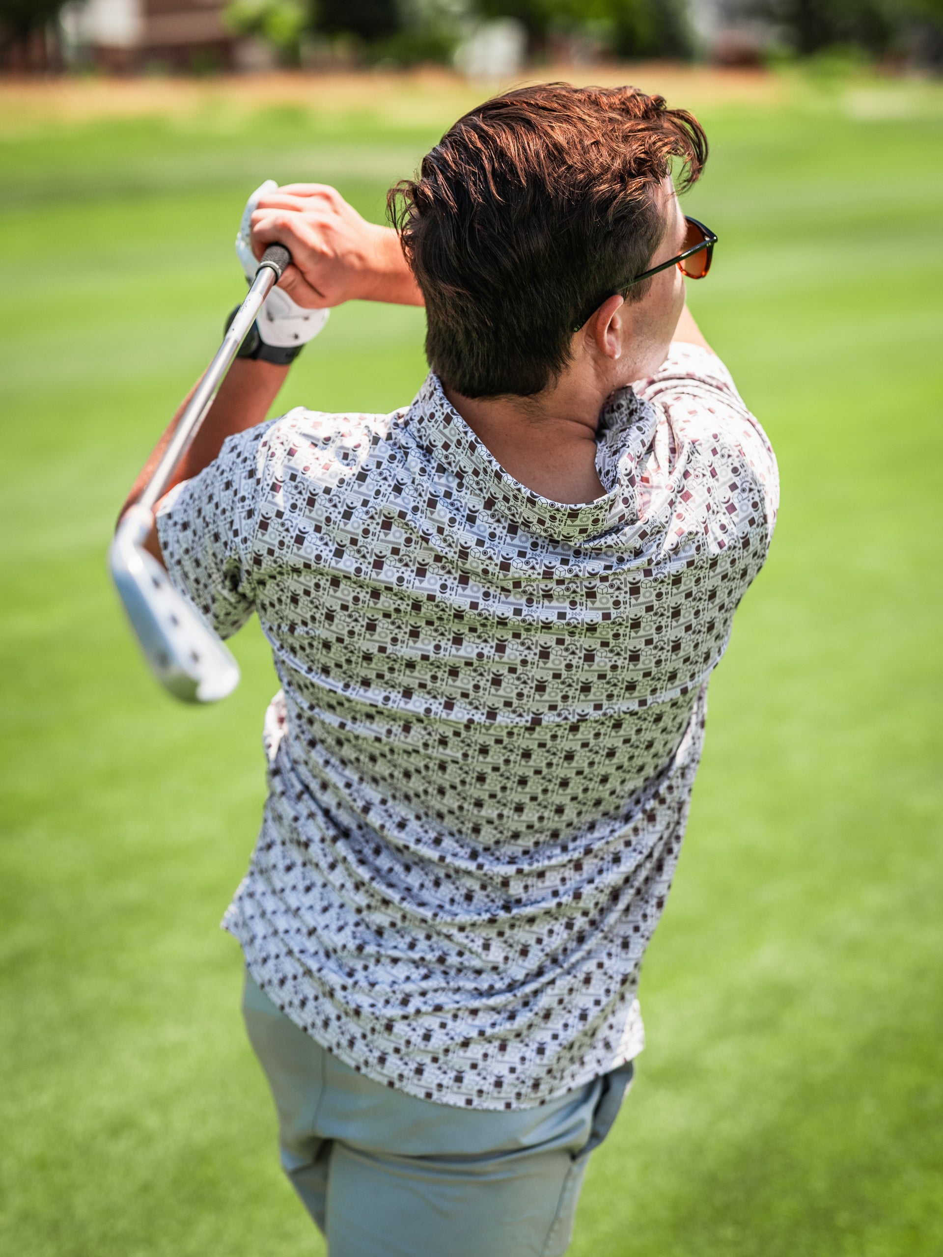 A golfer swings a club on the course, wearing a stylish retro-patterned shirt and sunglasses, showcasing bold golf fashion.