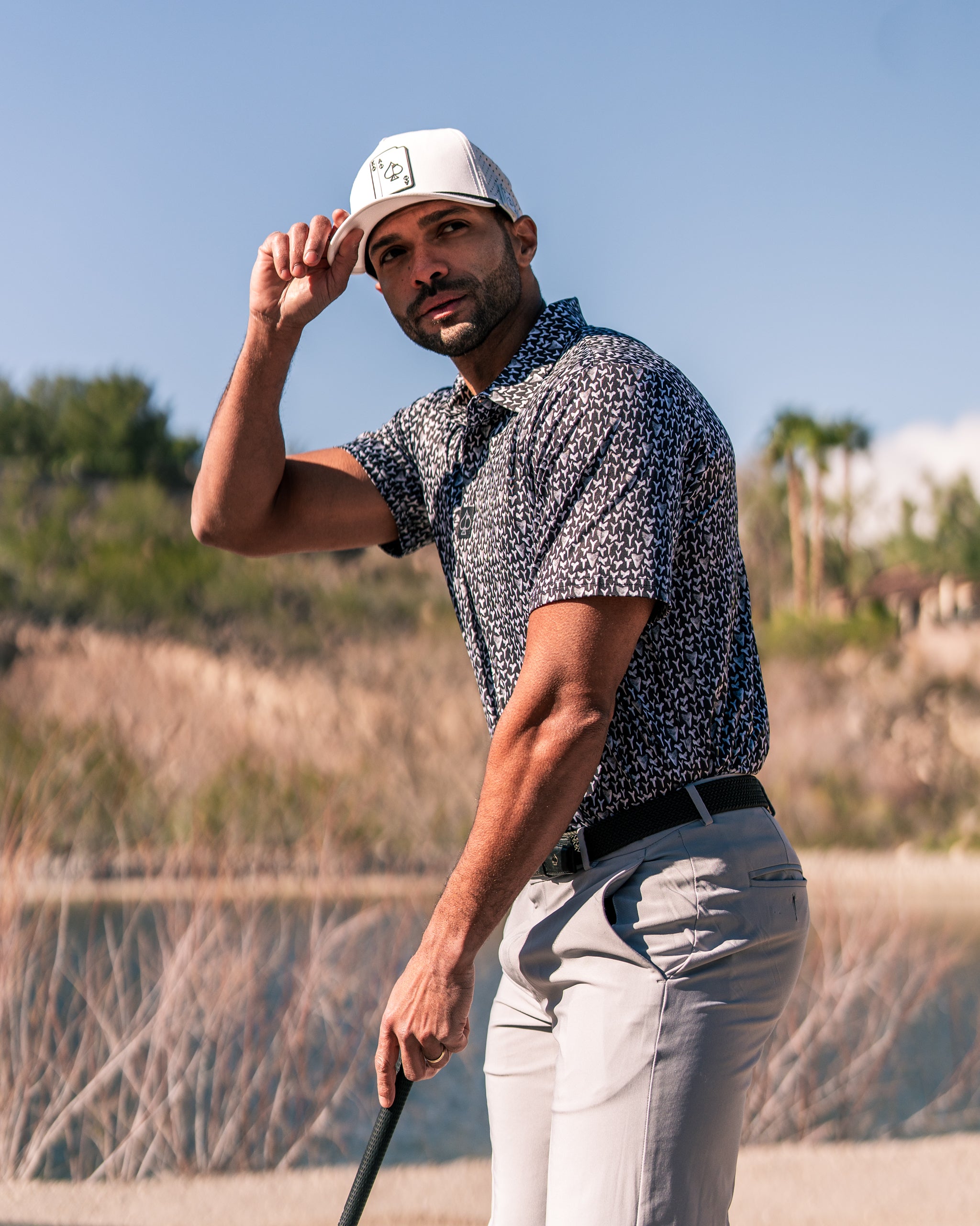 Man wearing a stylish Shark Tooth golf shirt and cap, posing confidently on a sunny golf course with a scenic background.