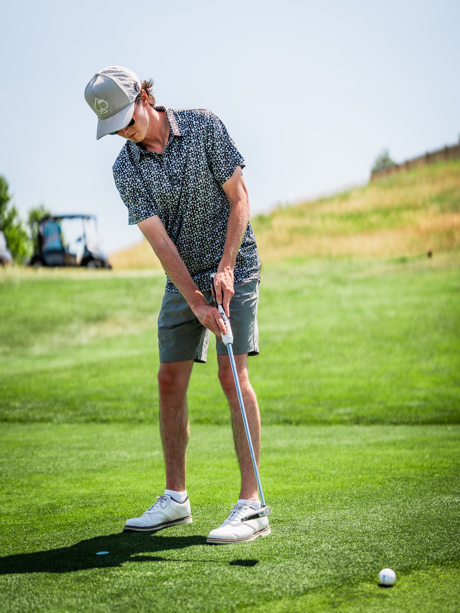 A golfer wearing a patterned shirt and cap prepares to putt on a sunny golf course, showcasing stylish golf apparel.