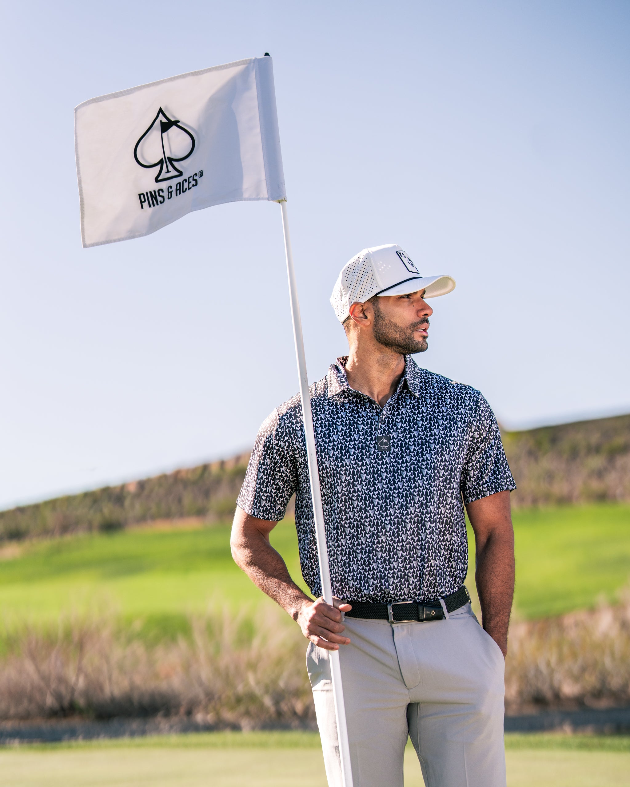 A golfer stands confidently by the flag on the course, wearing a stylish Shark Tooth shirt and cap from Pins and Aces.
